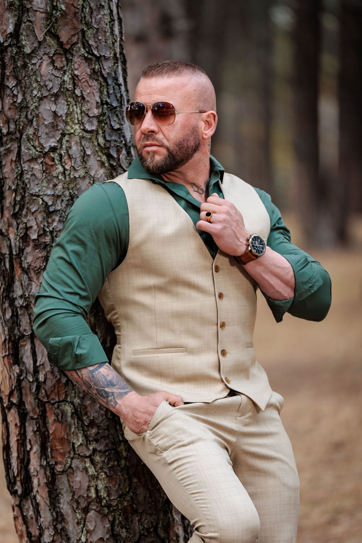 A man in a cream vest and green sleeves poses against a tree trunk in a forest setting.