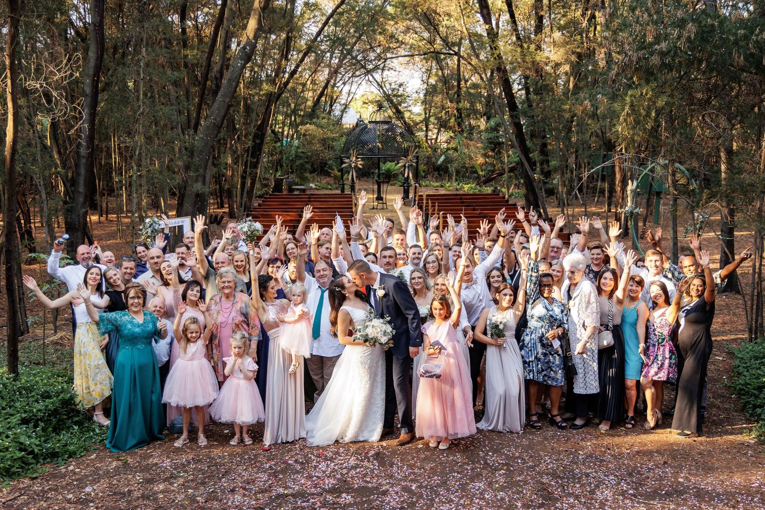 Wedding party celebrates outdoors with raised arms in a joyful group photo among trees.