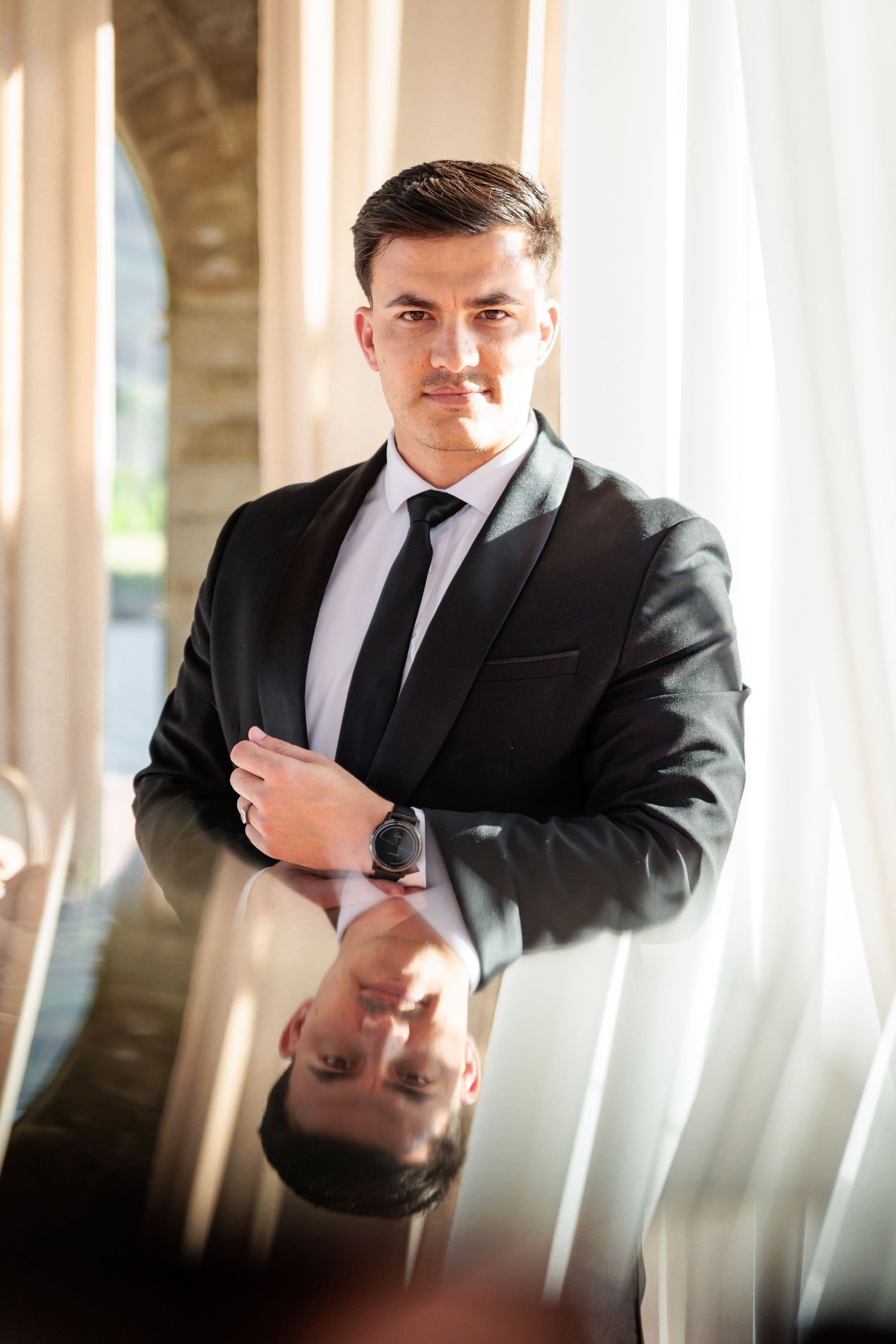 A person in a black suit and white tie adjusts their collar while standing in a room with vertical white pillars.