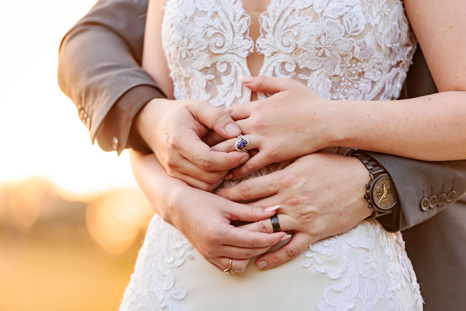 Close-up view of hands holding each other during a sunset wedding ceremony, with lace dress details visible in background.