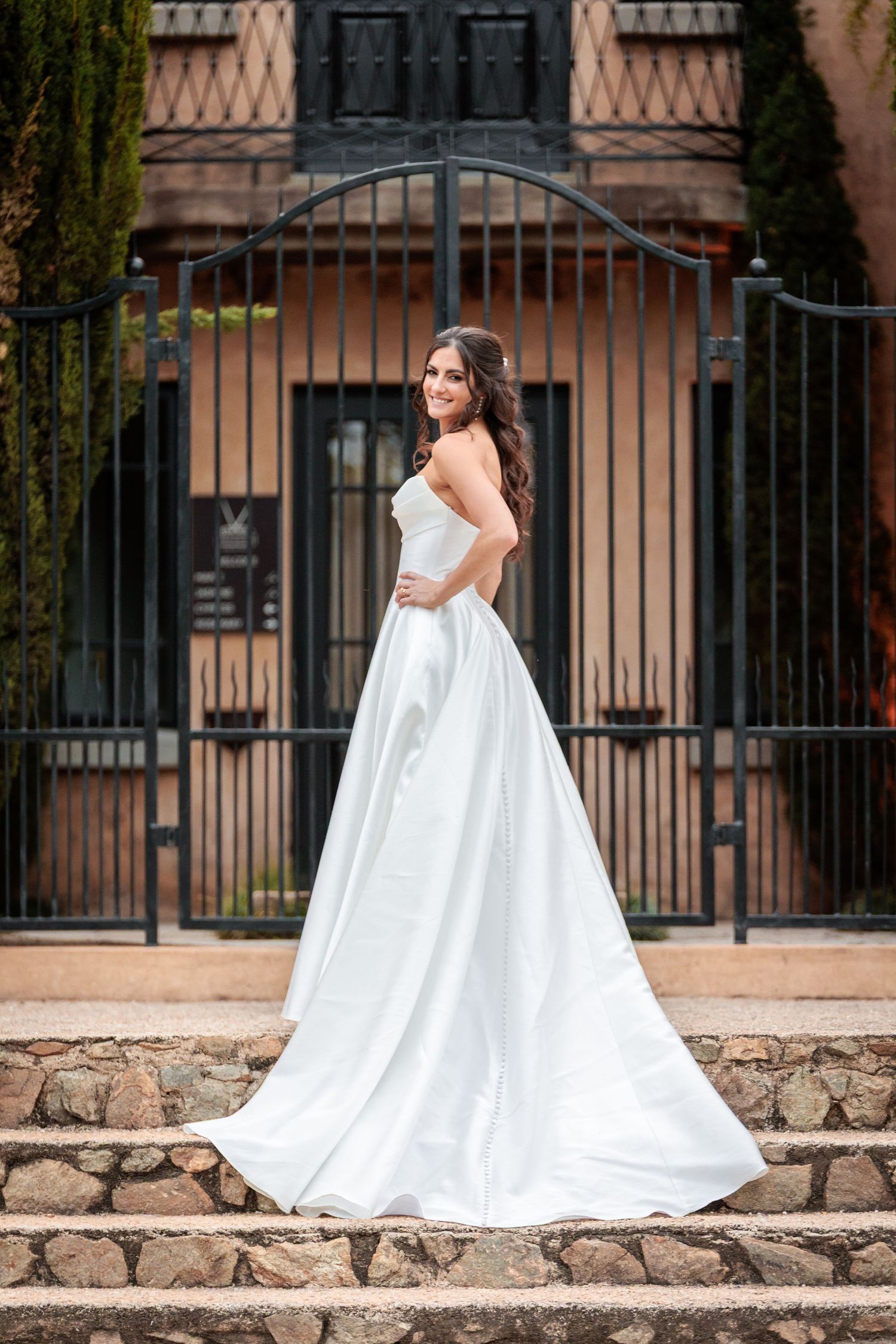A bride in a flowing white ballgown poses gracefully against a rustic stone wall and iron gate backdrop.