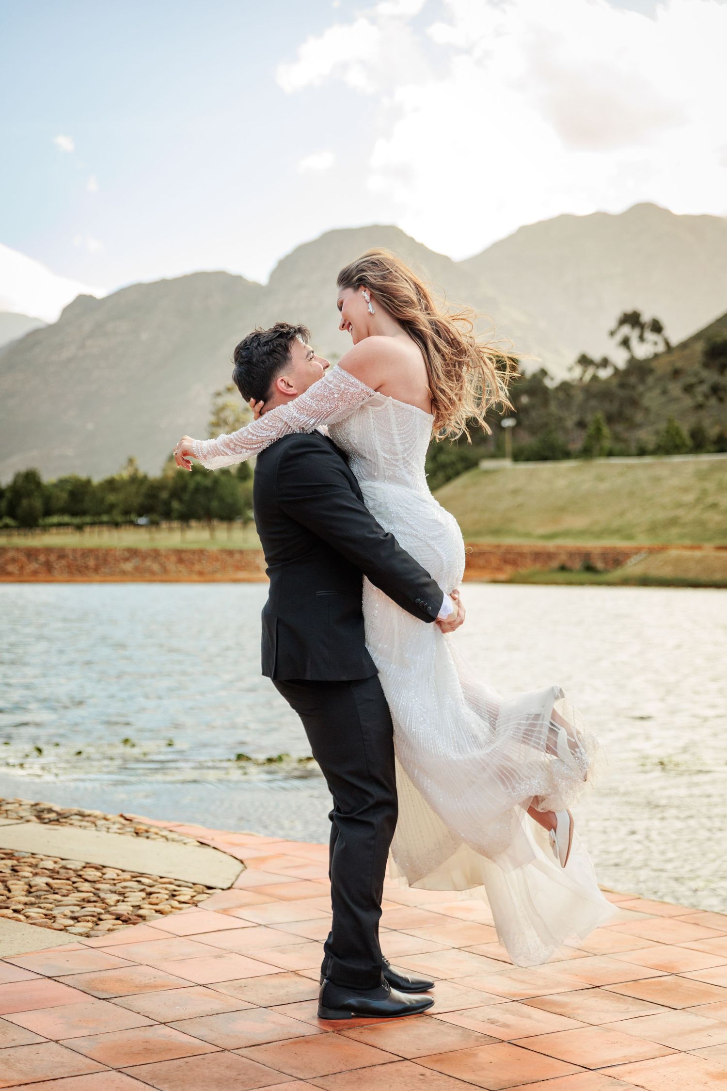 A romantic couple dances on a terrace overlooking a scenic mountain lake during a wedding celebration.
