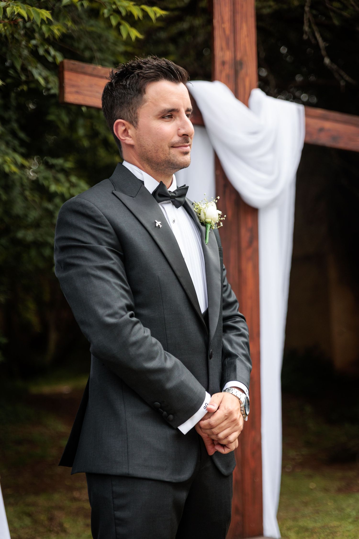 A groom in a black tuxedo stands at an outdoor wedding ceremony venue with wooden beams and white fabric decor.