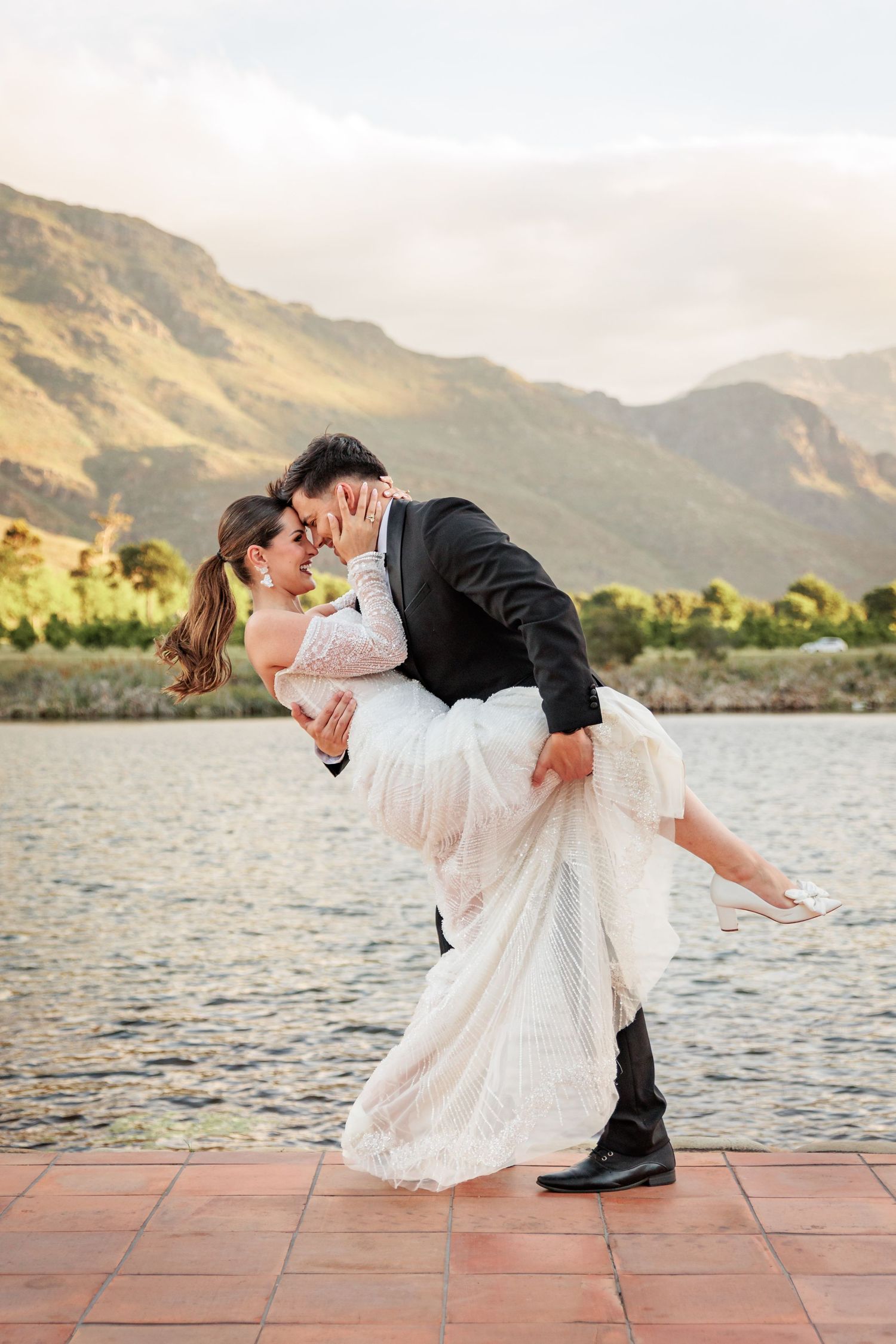 Romantic wedding couple shares a dramatic dip kiss against a scenic mountain lake backdrop at sunset.