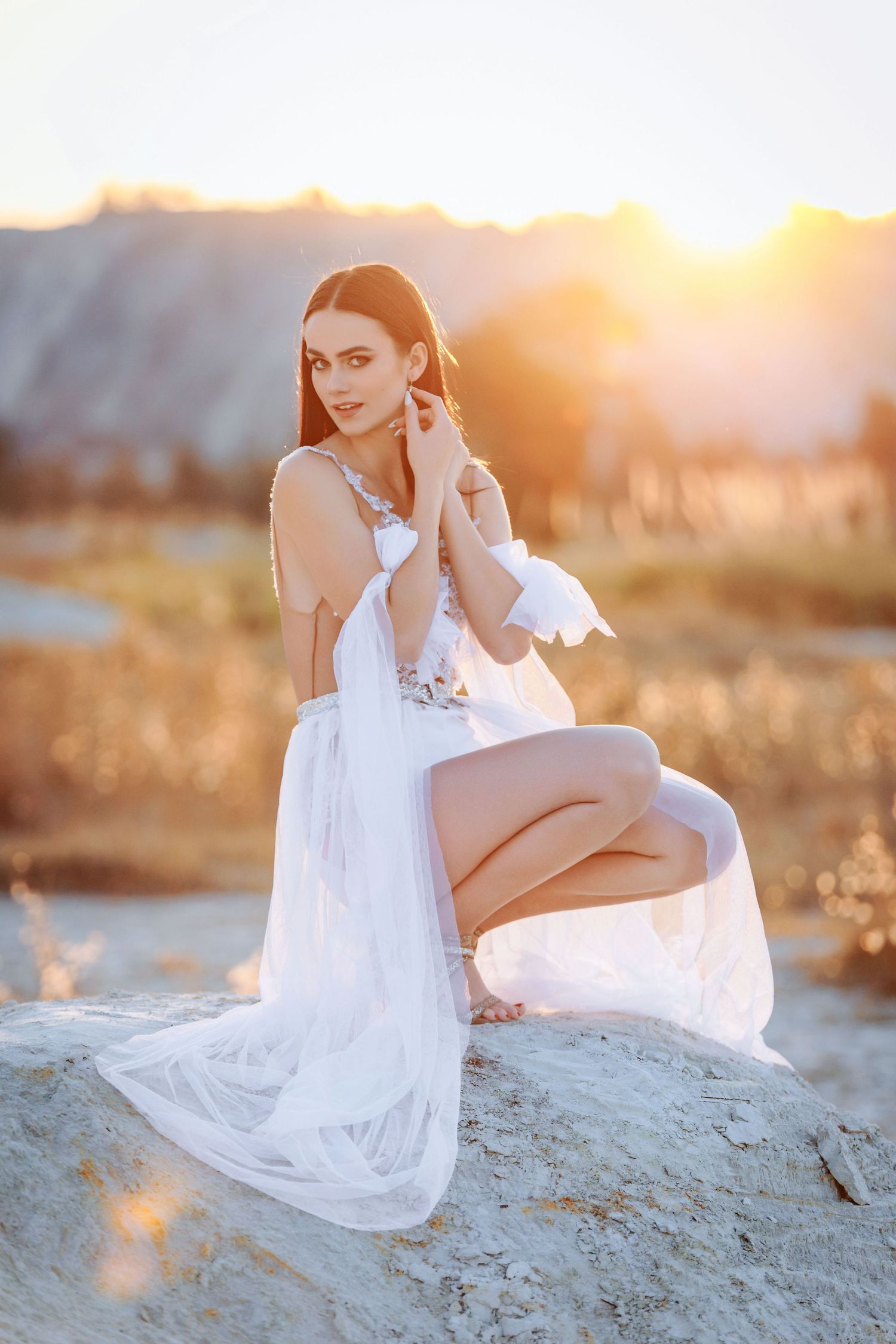 A figure in a flowing white dress poses gracefully on rocks during golden hour sunset.