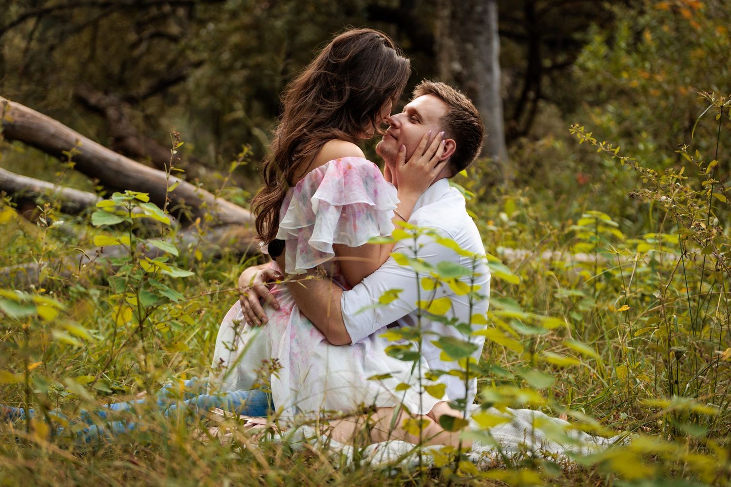 Two people in white outfits embrace tenderly on grassy ground surrounded by nature and foliage.