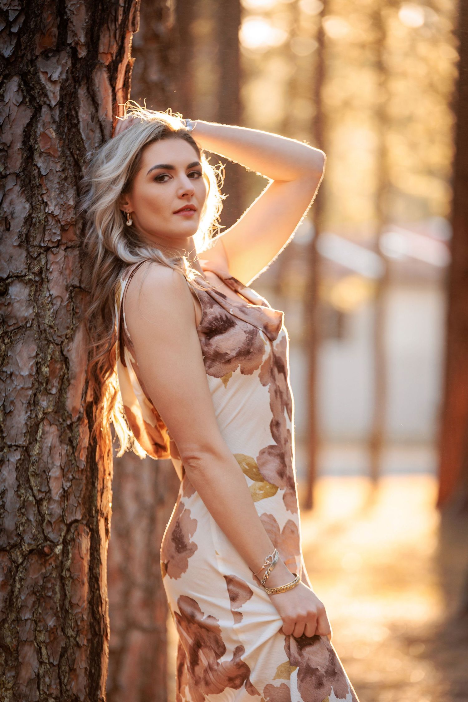 A woman in a floral dress poses against a tree trunk at sunset in a forest with warm golden lighting.