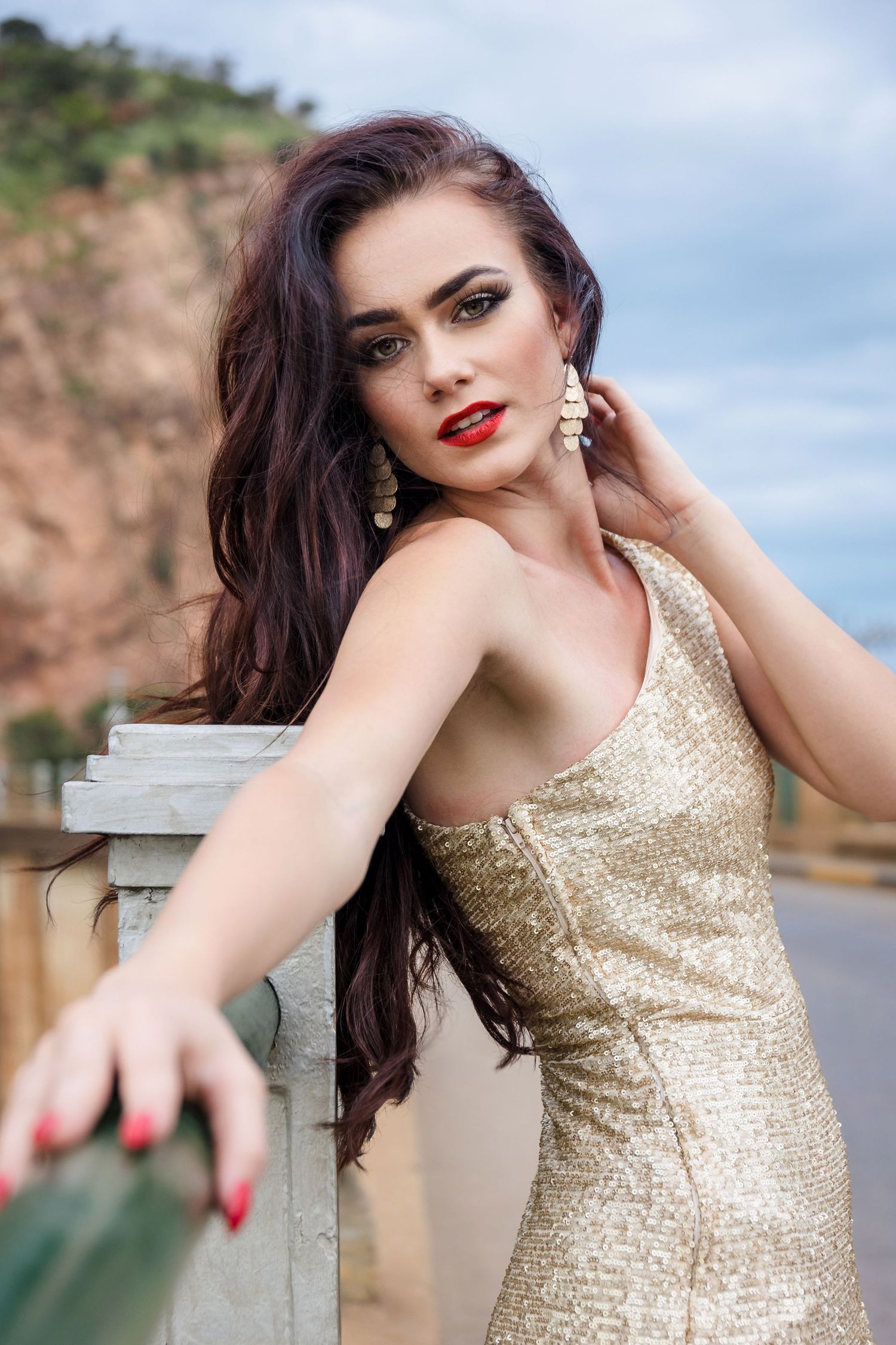 A woman in a sparkling gold dress poses elegantly against a scenic outdoor backdrop.