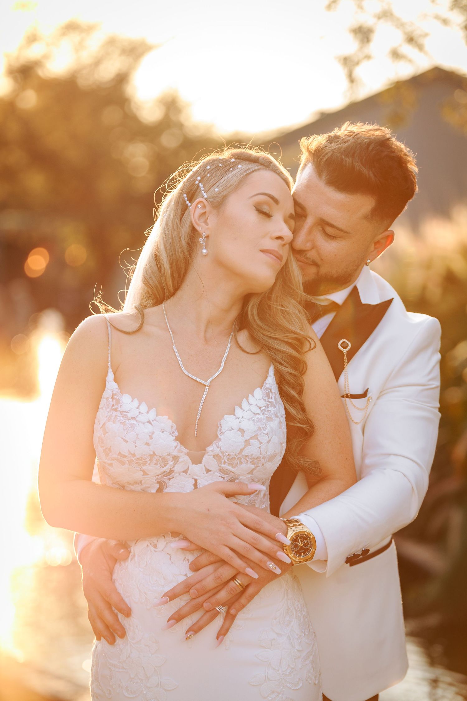Romantic wedding couple embraces in golden sunset light wearing white formal attire and lace gown.
