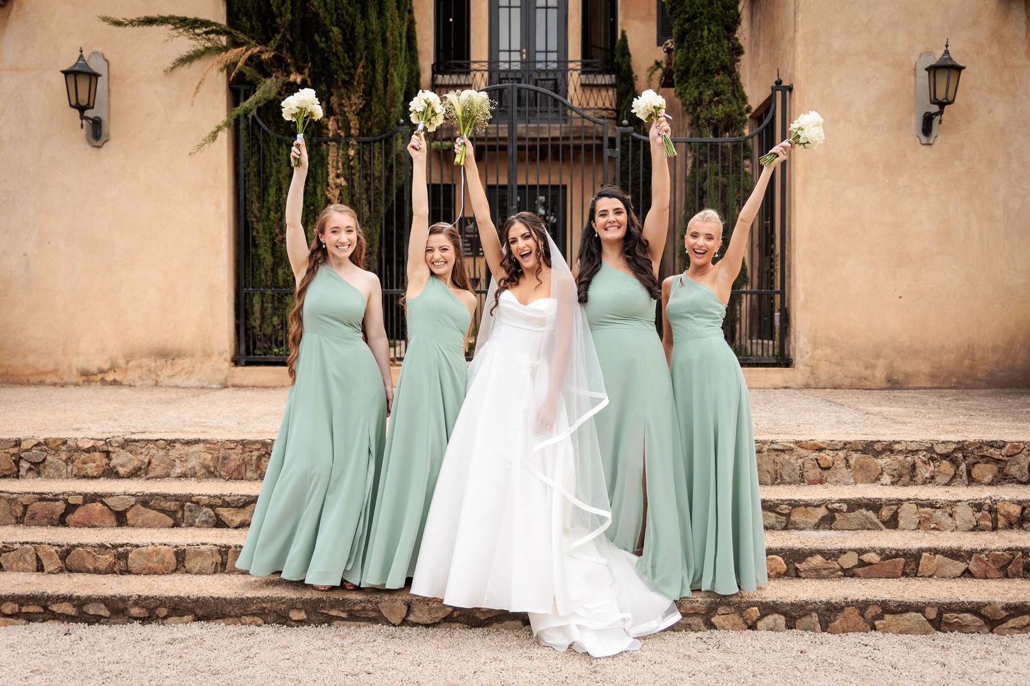 Wedding party in mint green bridesmaid dresses and white bridal gown celebrating on stone steps of Mediterranean venue.