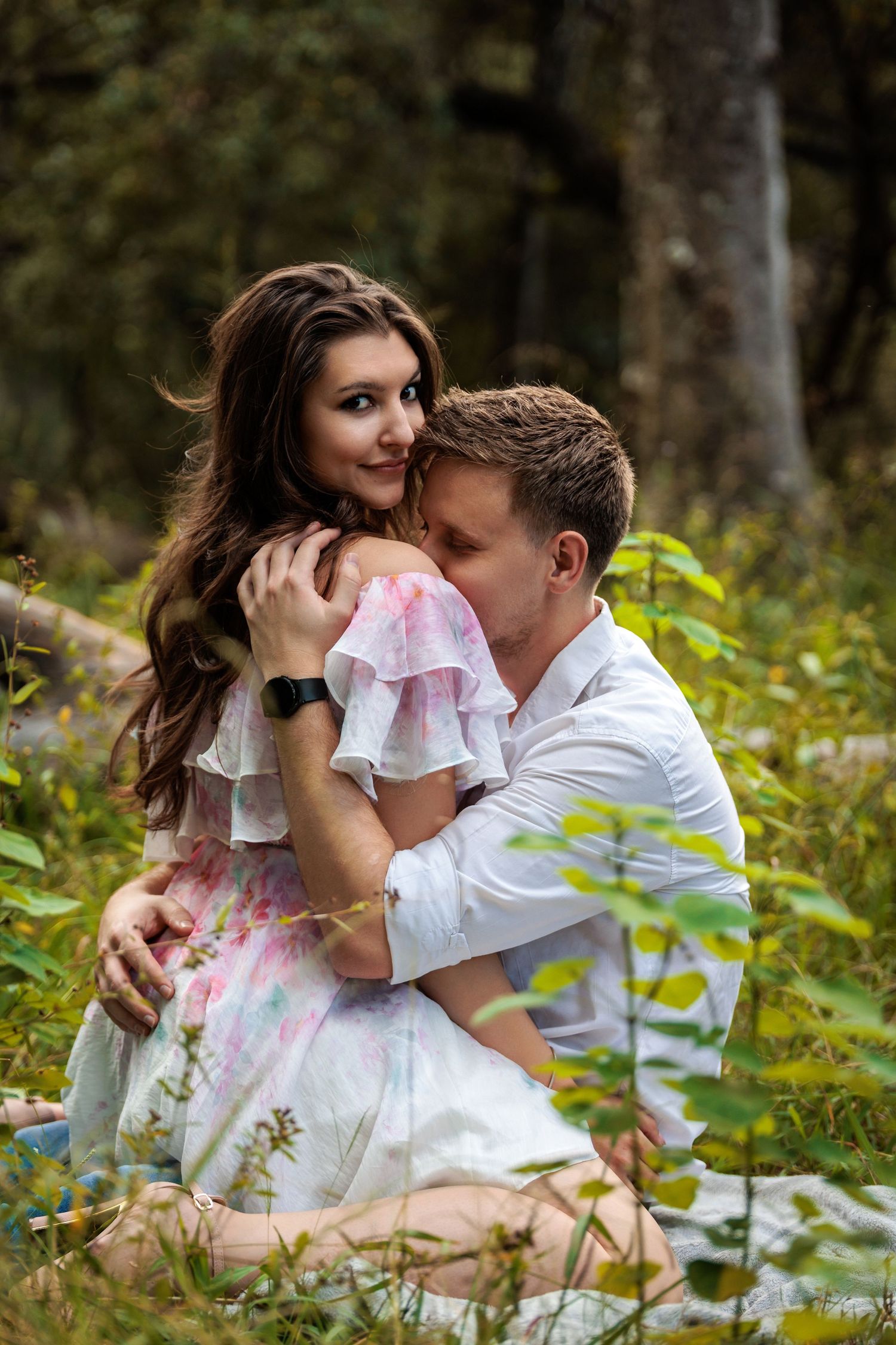 A romantic couple embraces in a natural outdoor setting surrounded by lush greenery and wildflowers.
