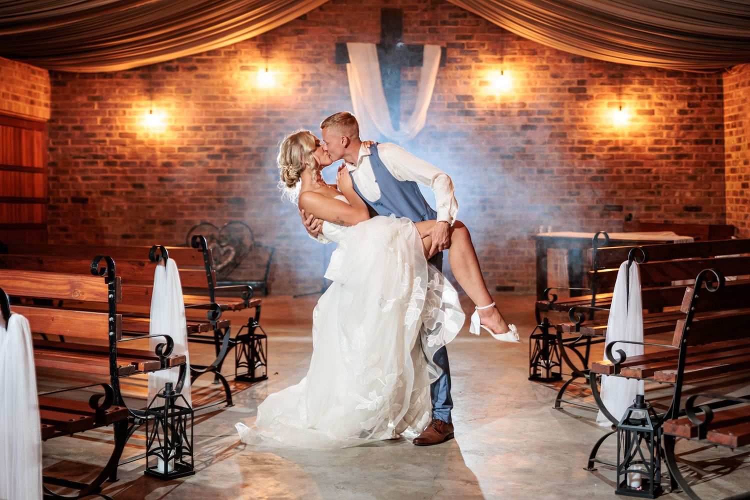 Bride in flowing white gown poses in rustic brick venue with vintage sewing machines and warm lighting.