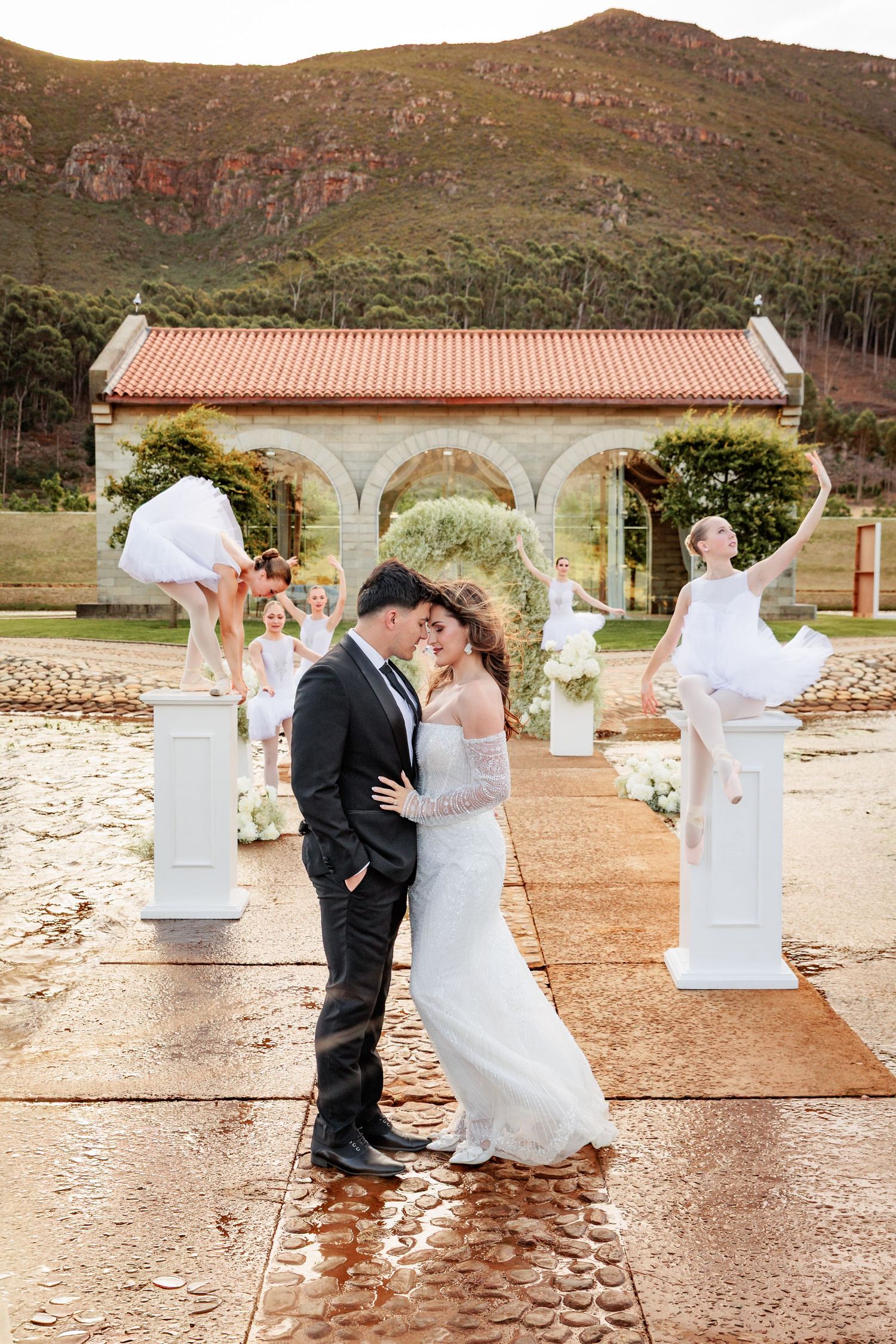 A couple shares romantic moments at a rustic outdoor venue with stone columns and red tile roofs in the background.