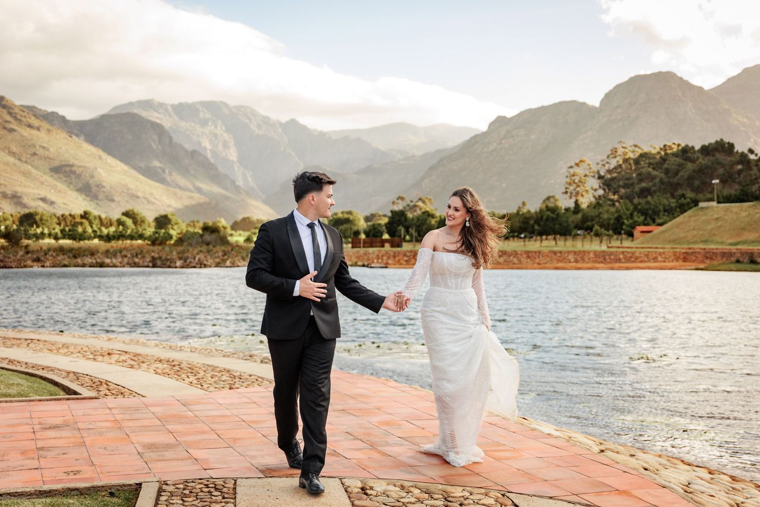 Bride and groom holding hands walking by a lake with stunning mountain backdrop on their wedding day.