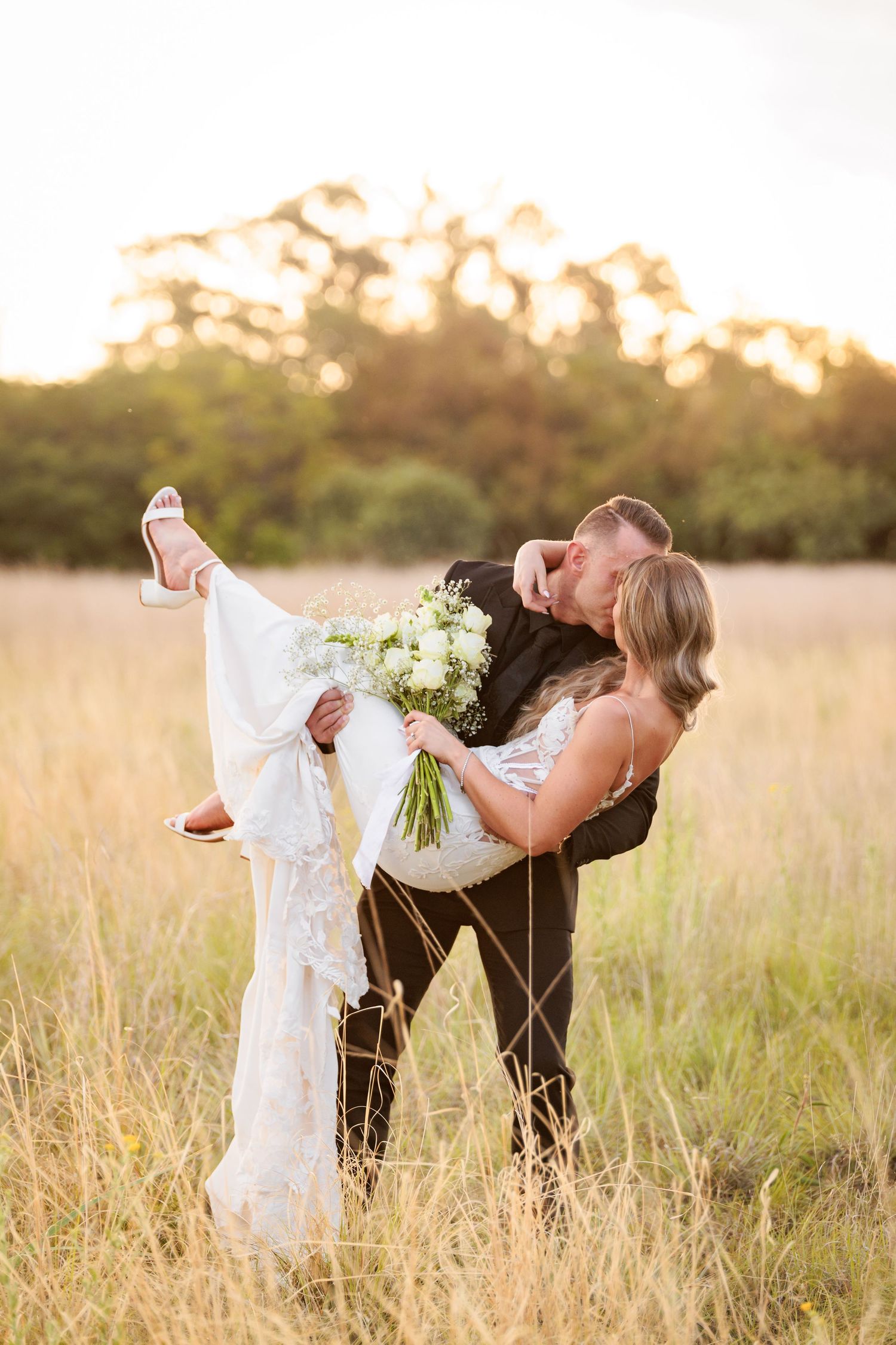 A romantic couple embraces in a golden field at sunset while holding a bouquet of white flowers.