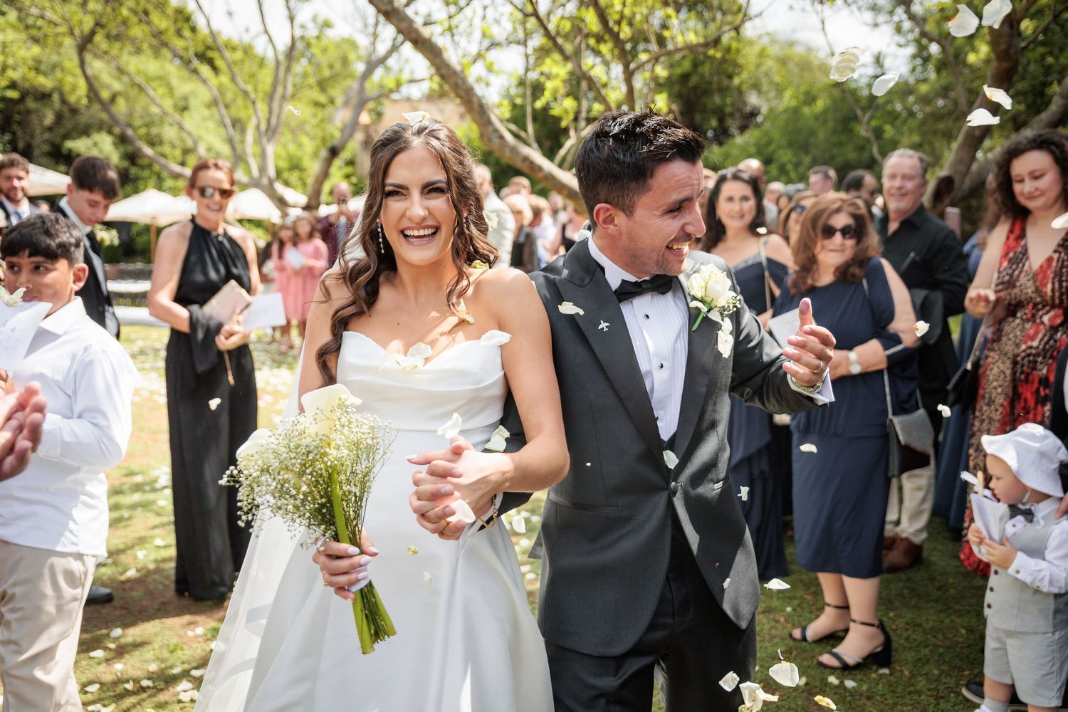 Newlyweds walk through crowd of guests throwing confetti at outdoor garden wedding ceremony.