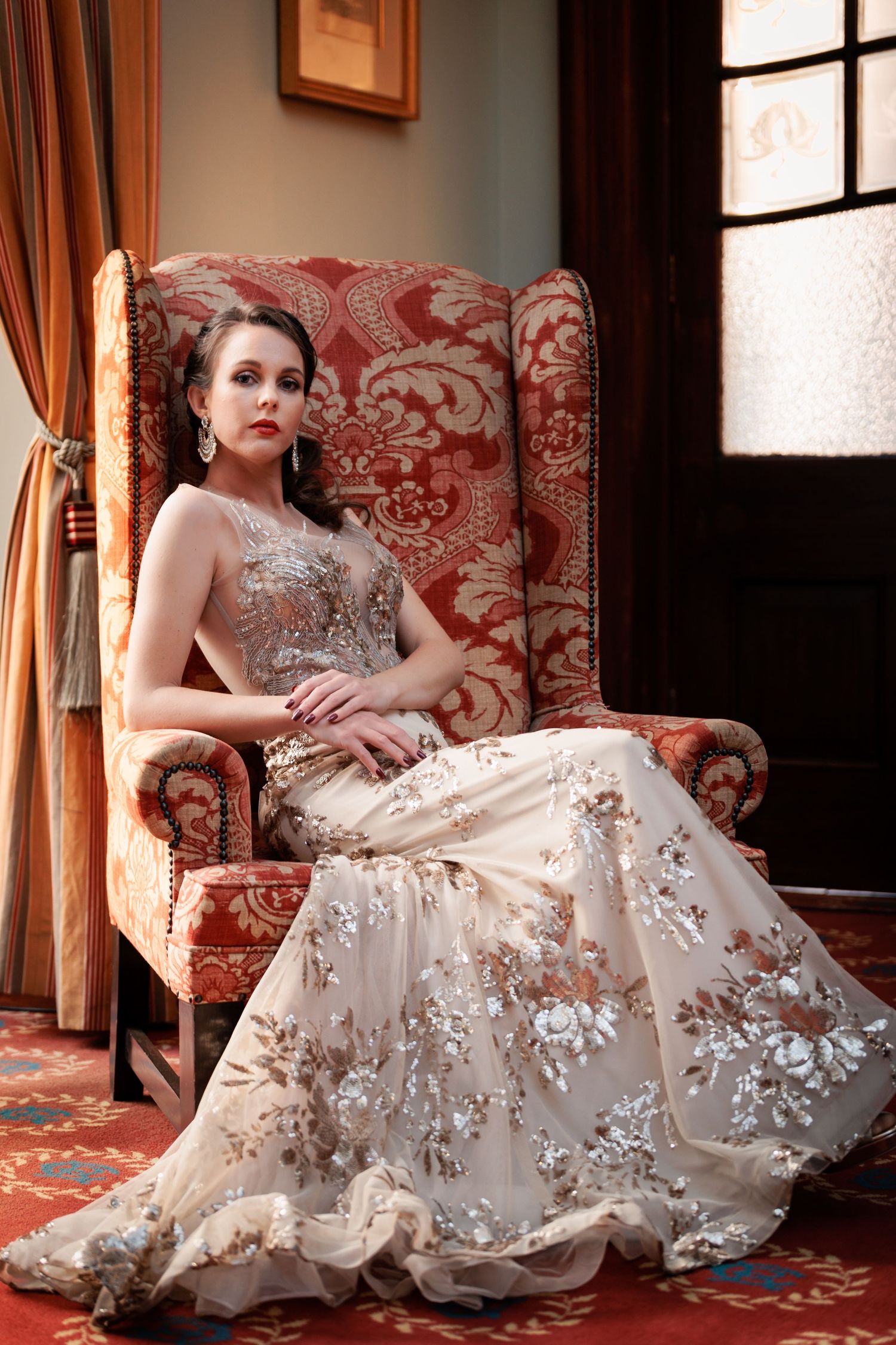 Elegant figure in a cream lace gown with floral appliques poses gracefully on a red damask chair in a luxurious setting.