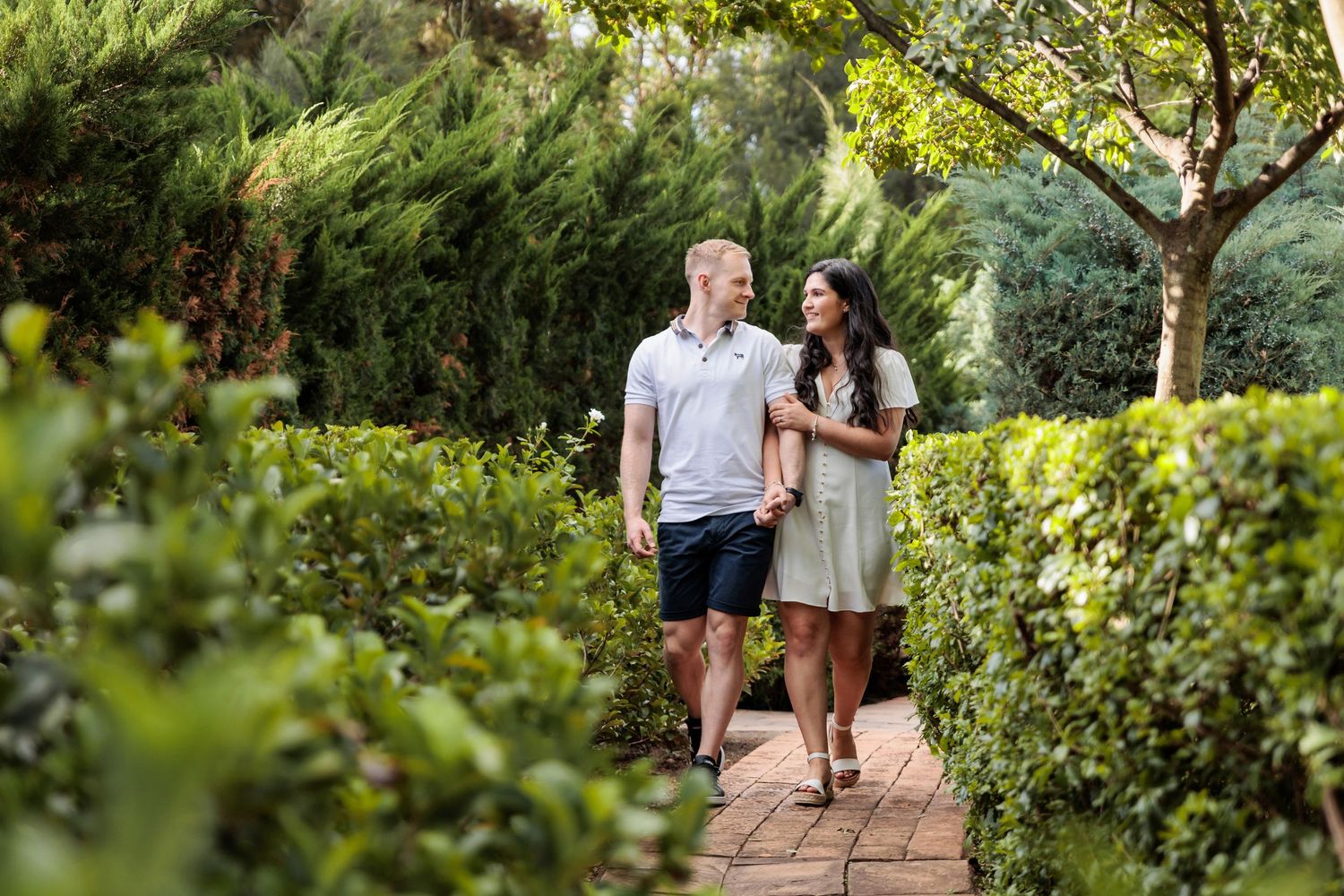 A couple walks together on a garden path surrounded by lush greenery and trees on a sunny day.