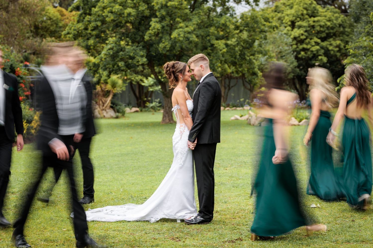 A wedding party in green dresses and black suits poses together on a lush green lawn during an outdoor celebration.