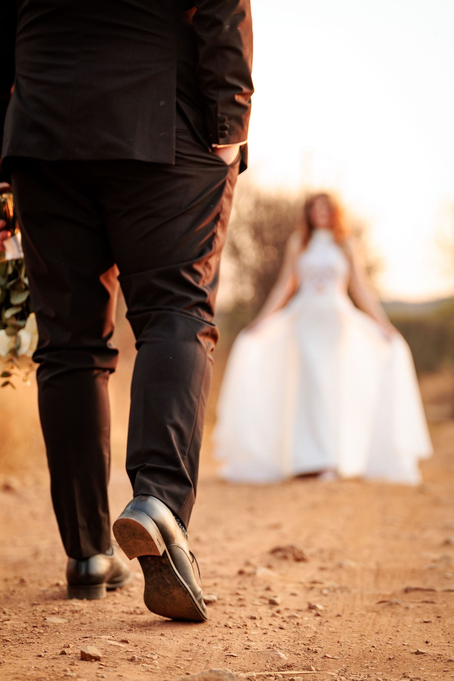 A groom walks on a dirt path towards his bride in a white wedding dress during golden hour sunset.