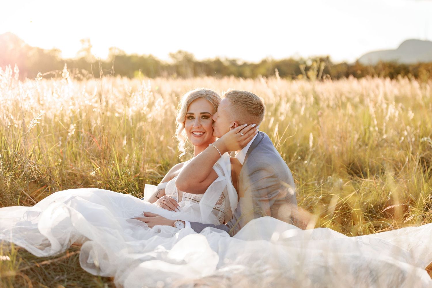 A couple sits together in a sun-drenched field of tall golden grass during sunset.