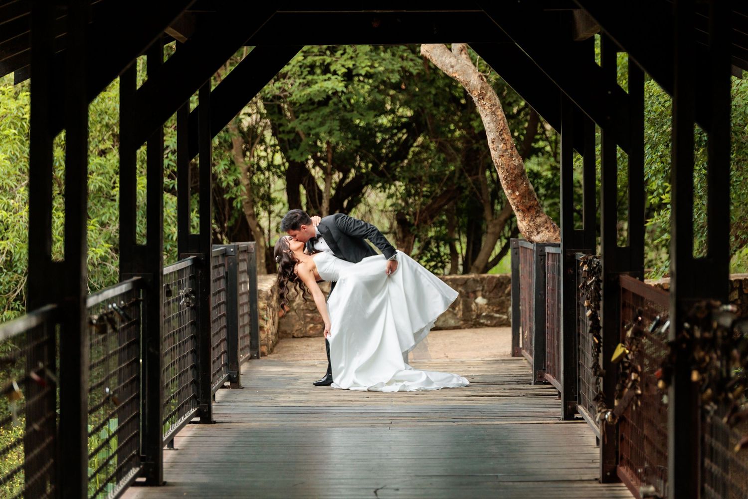 A bride and groom share a romantic moment under a tree-lined pathway in a lush garden setting.