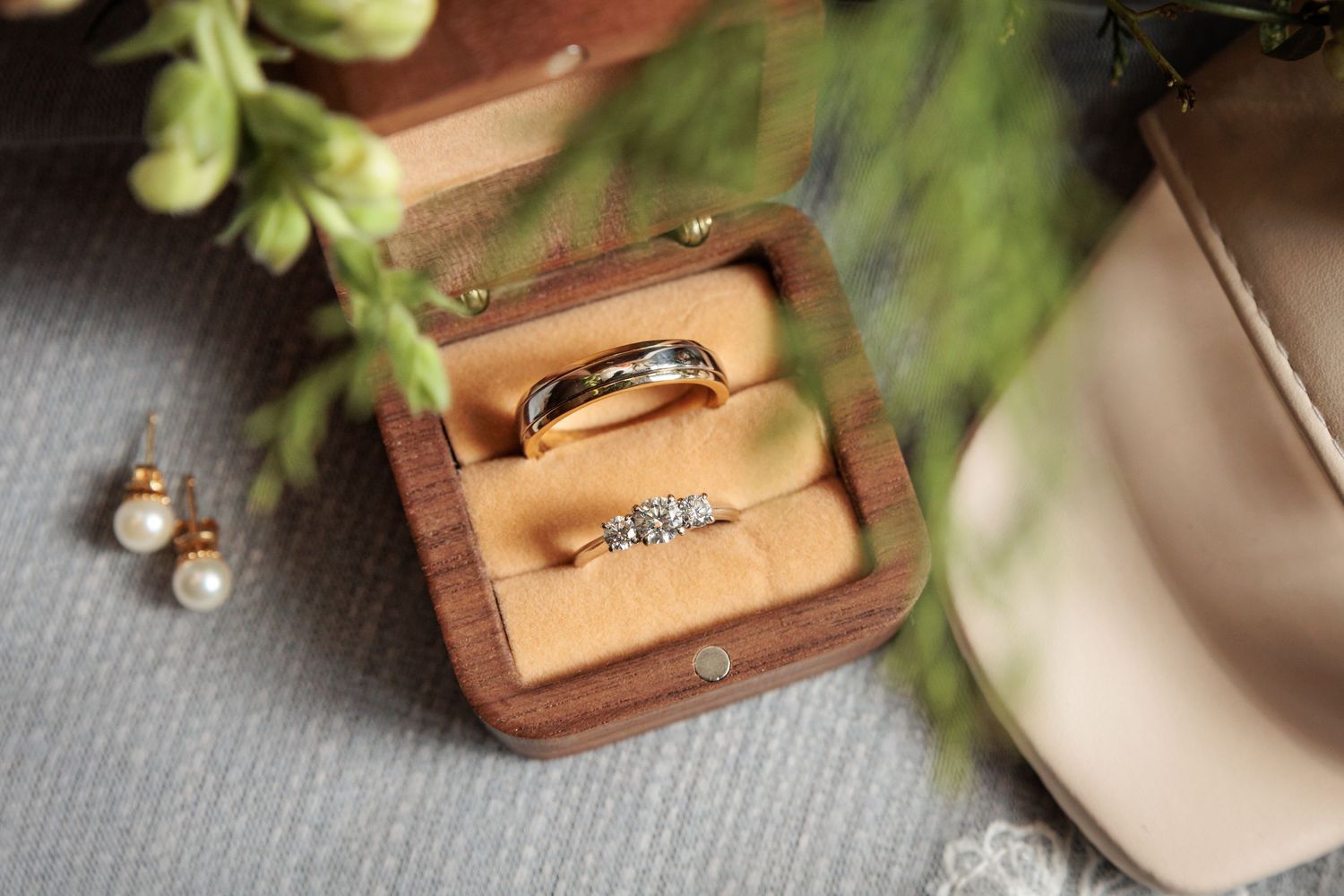 Gold wedding band and diamond engagement ring displayed in a wooden ring box with pearl earrings nearby.