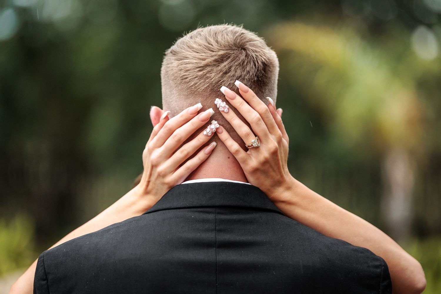 Bride places hands on groom's neck from behind, showing engagement ring, with green trees in background.