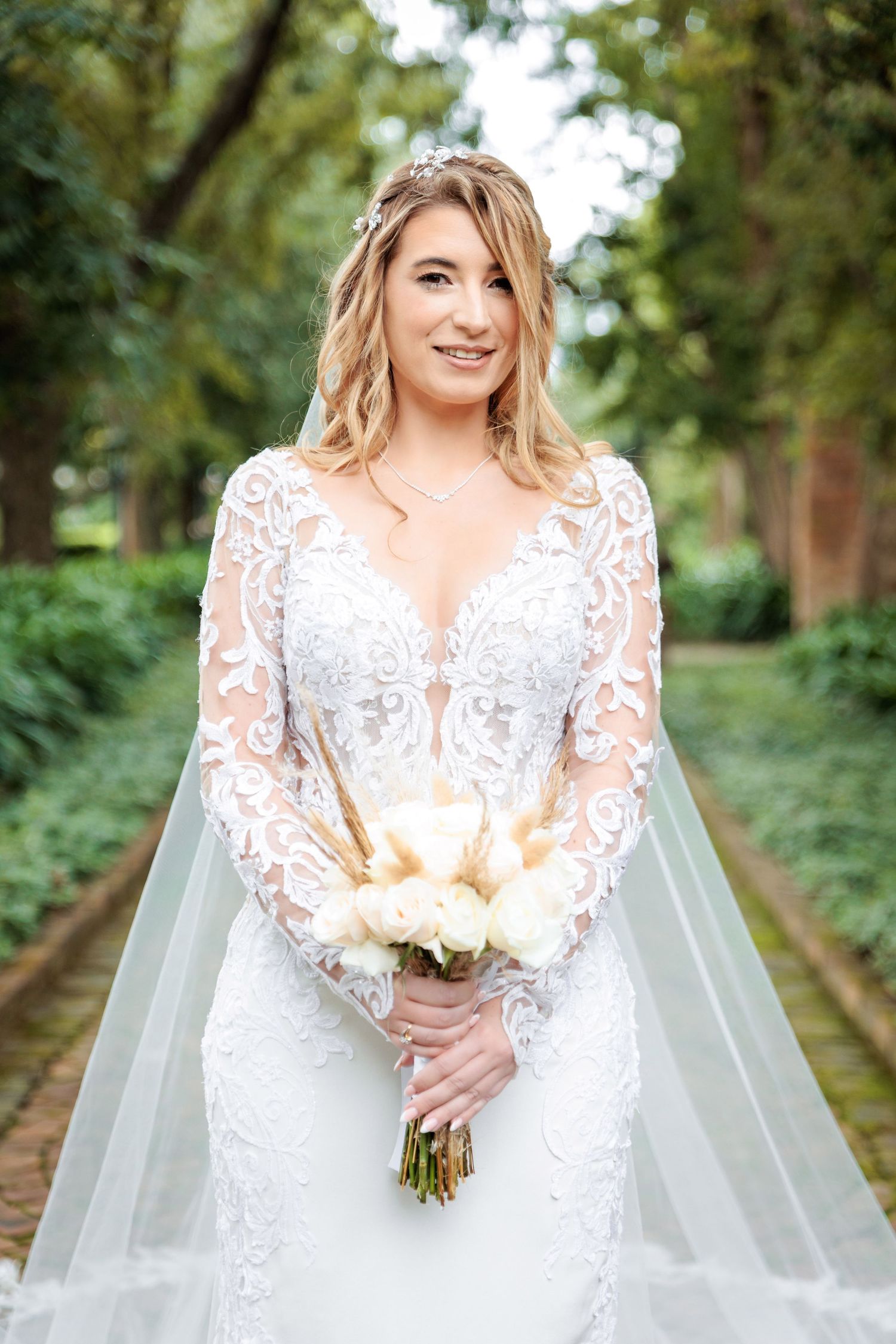 Bride in long sleeve lace gown holds white bouquet on garden path.