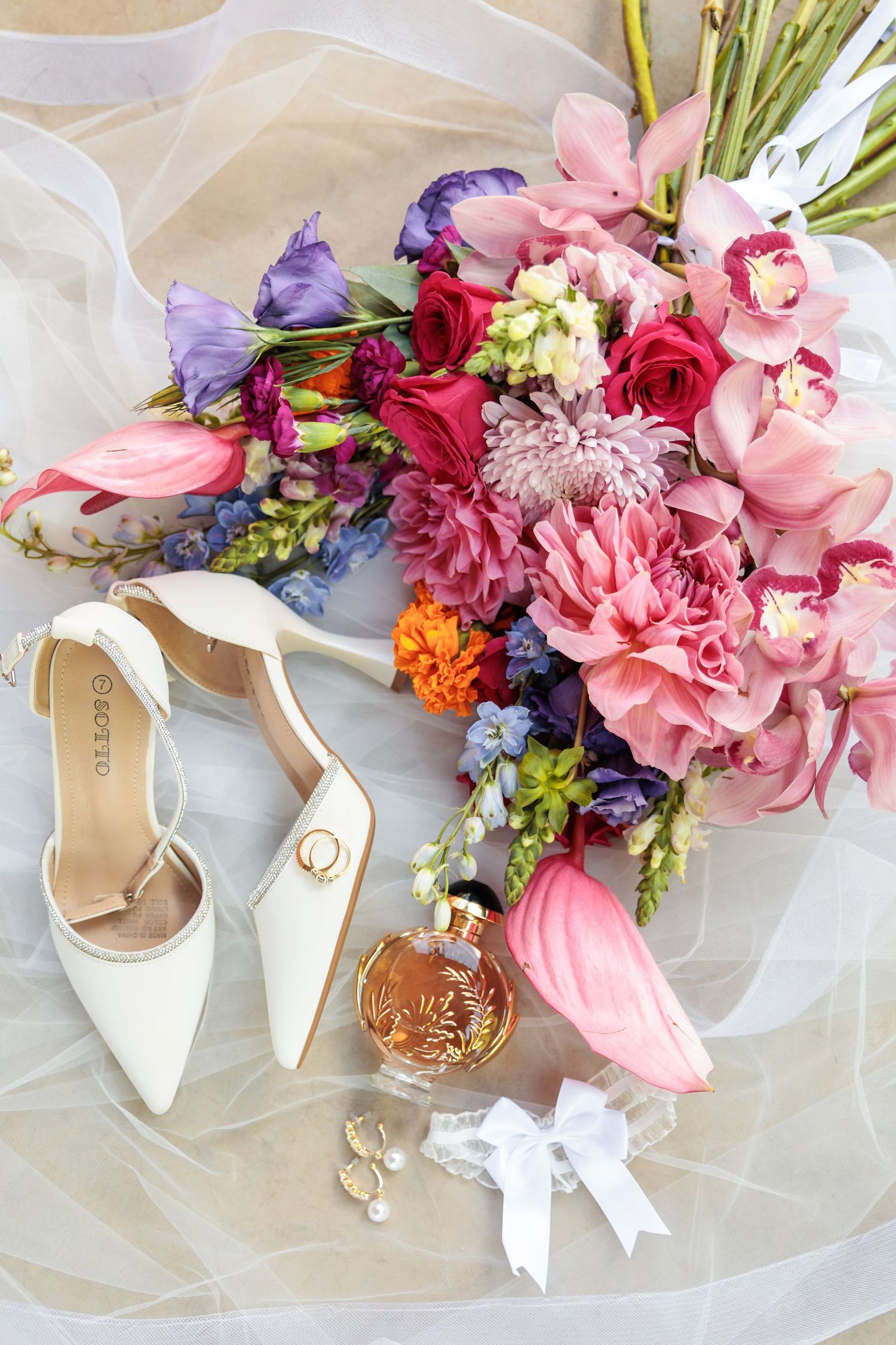 Elegant wedding flatlay with white bridal shoes and vibrant pink and purple floral bouquet on sheer fabric.
