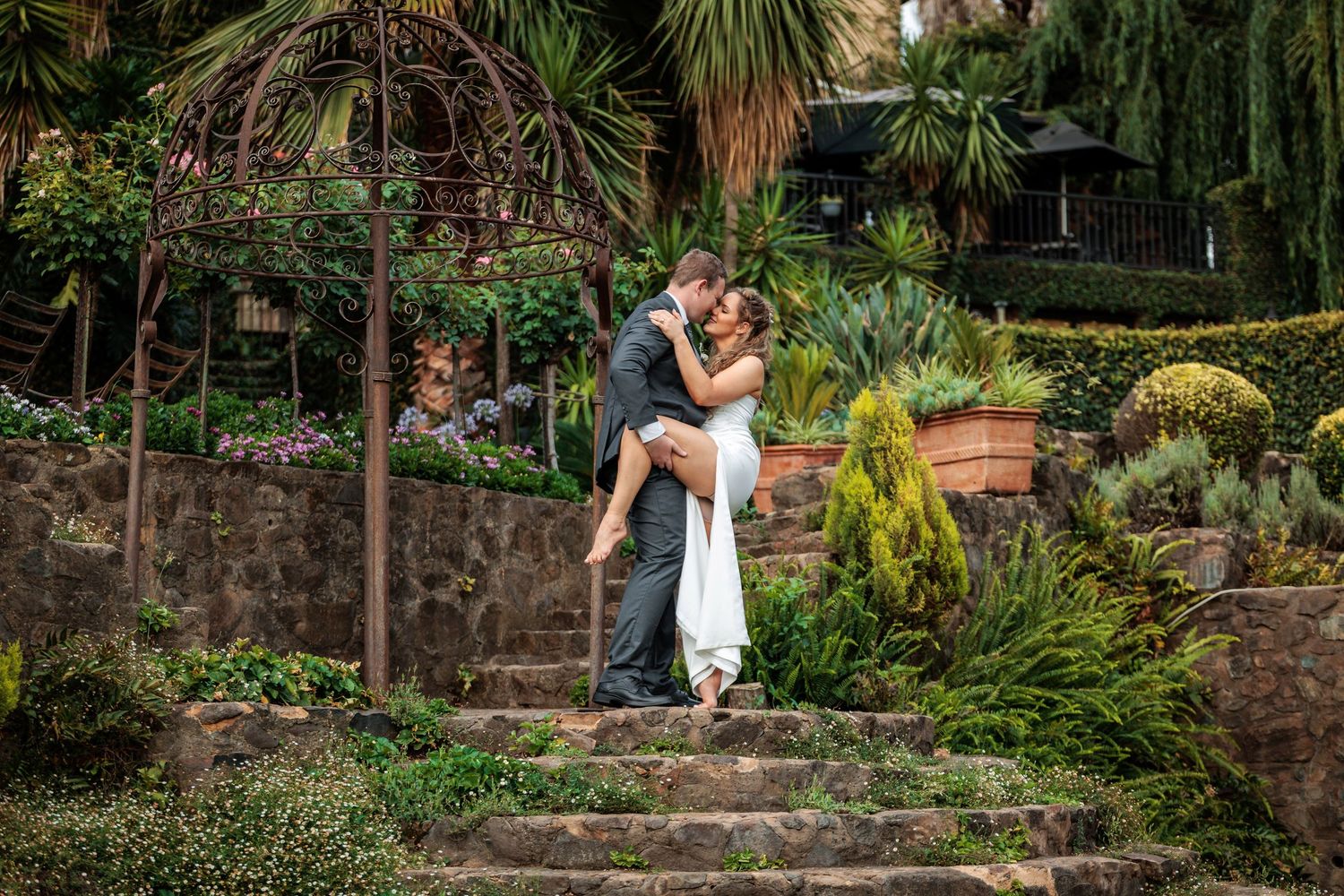 A couple shares an intimate moment on stone steps surrounded by tropical gardens and palm trees.
