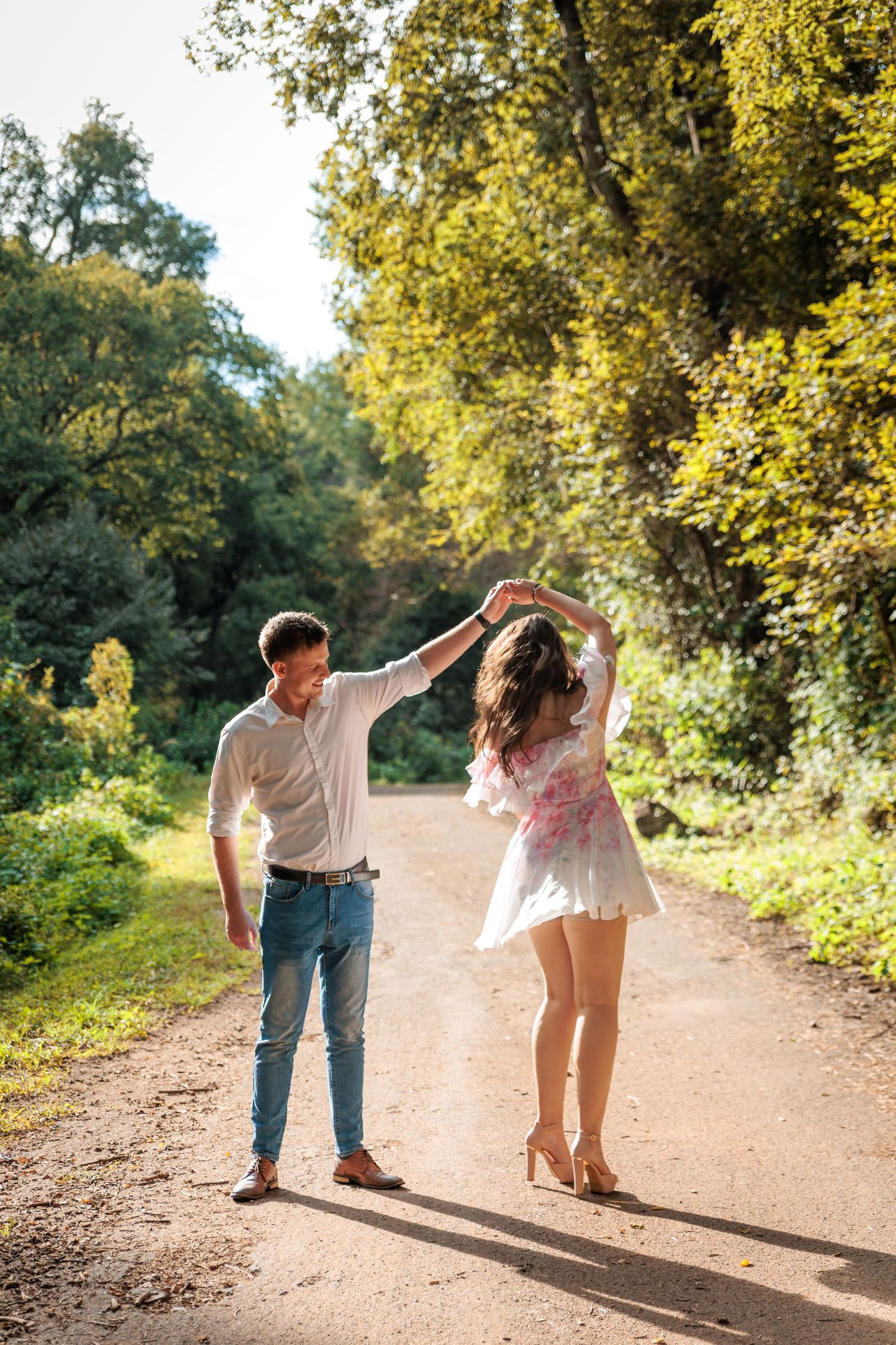 A couple dancing playfully together on a sunlit dirt path surrounded by lush green and yellow foliage.