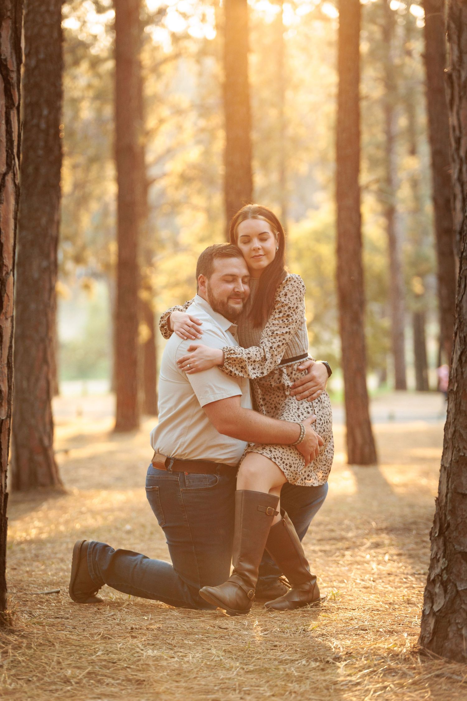 A couple sits together in a sunlit forest with golden autumn light filtering through tall pine trees.