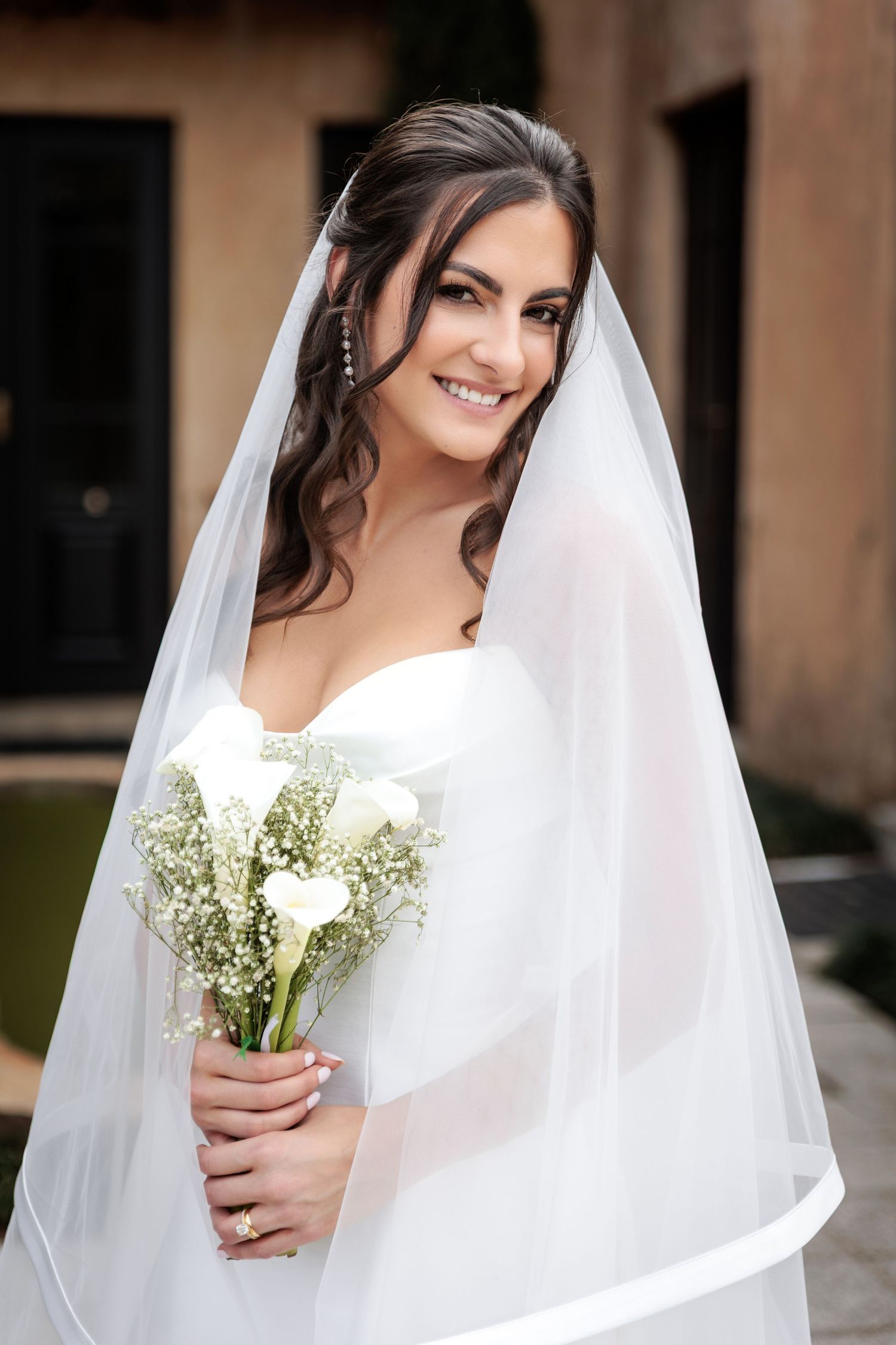 A bride in a white wedding dress and veil holds a delicate bouquet of baby's breath flowers while smiling.