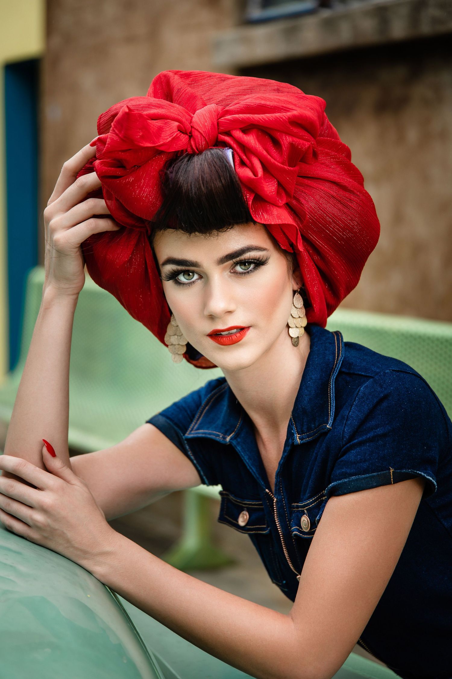 Retro-inspired fashion portrait featuring a dramatic red headpiece and dark denim jacket against a teal background.