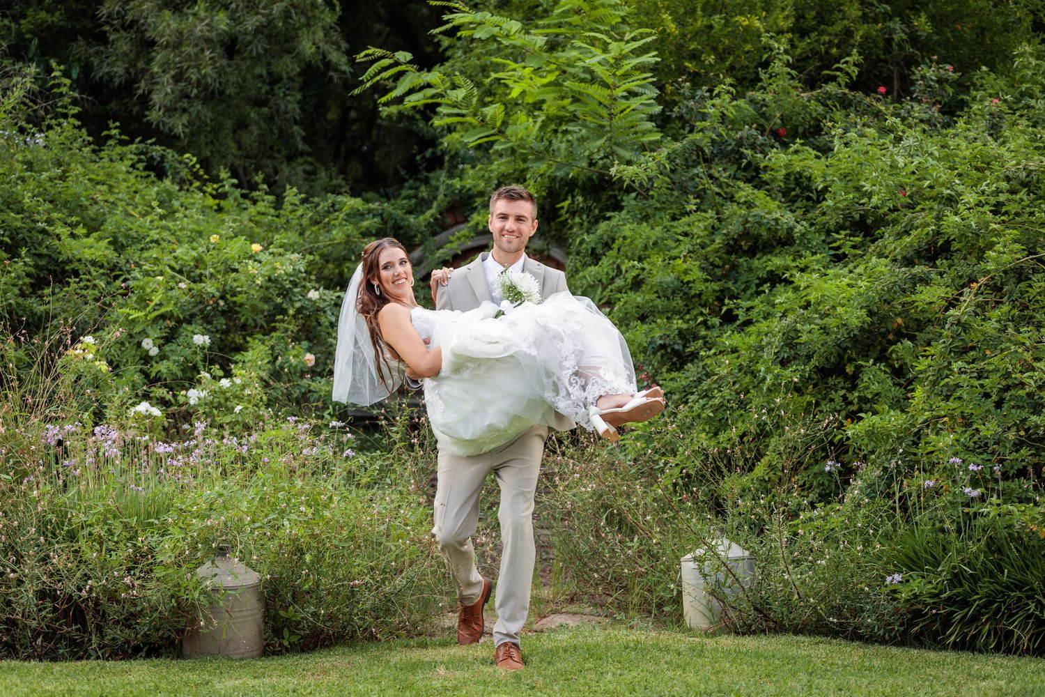 Playful wedding couple dances and spins outdoors among green foliage.