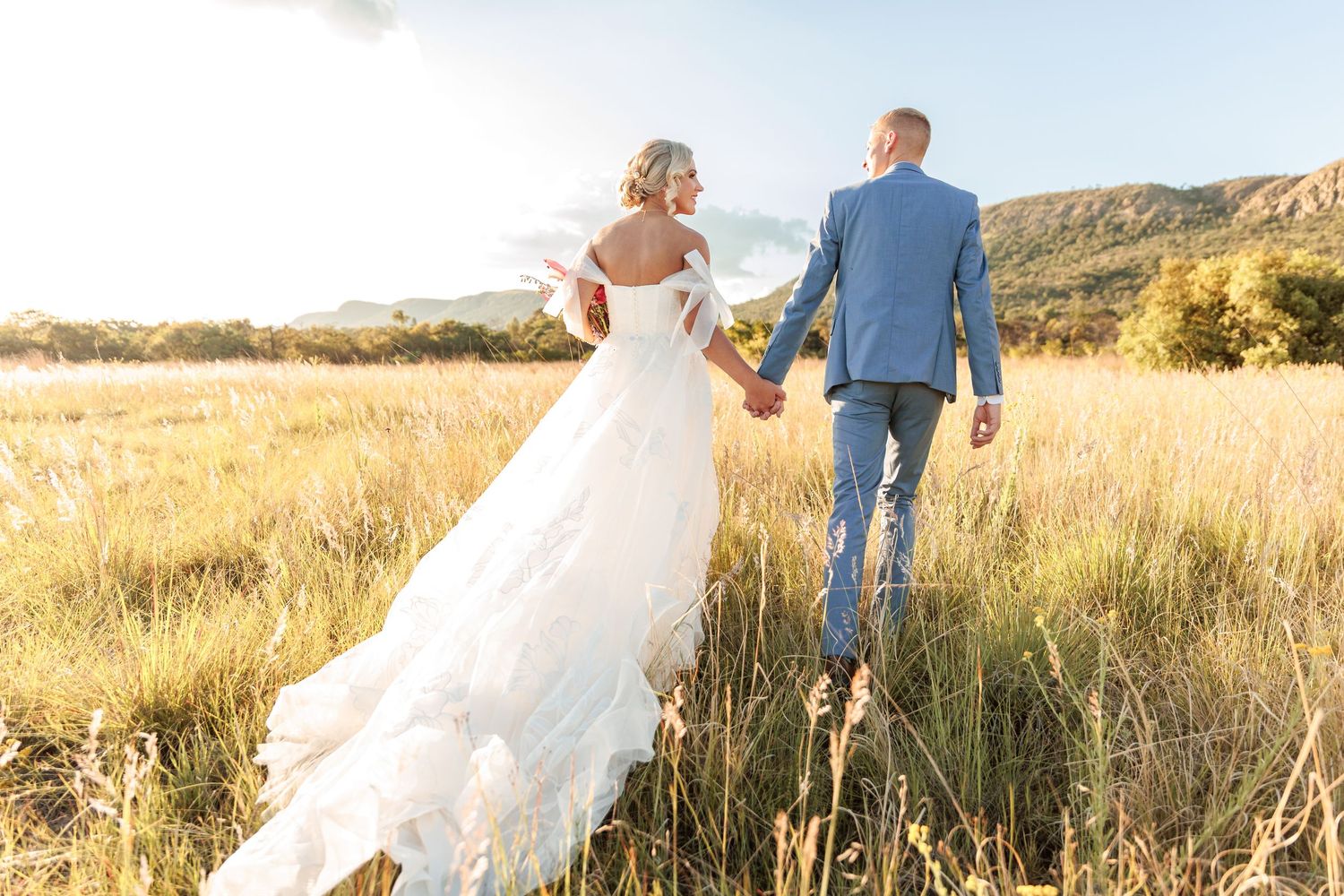 A couple in wedding attire walks through a golden field at sunset with mountains in the background.