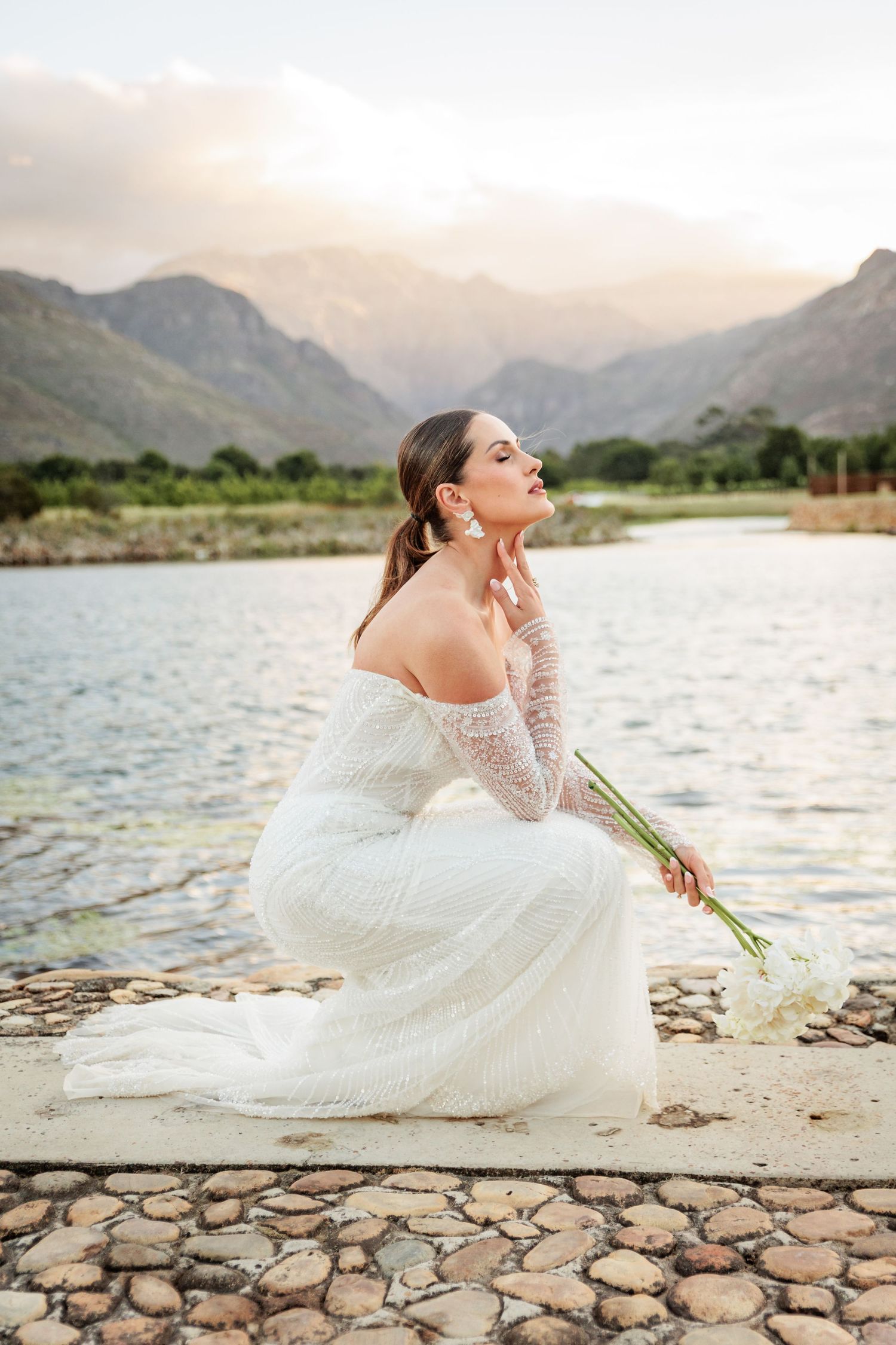 A bride in a white lace gown reclines on a stone wall by a lake with mountains in the background at sunset.