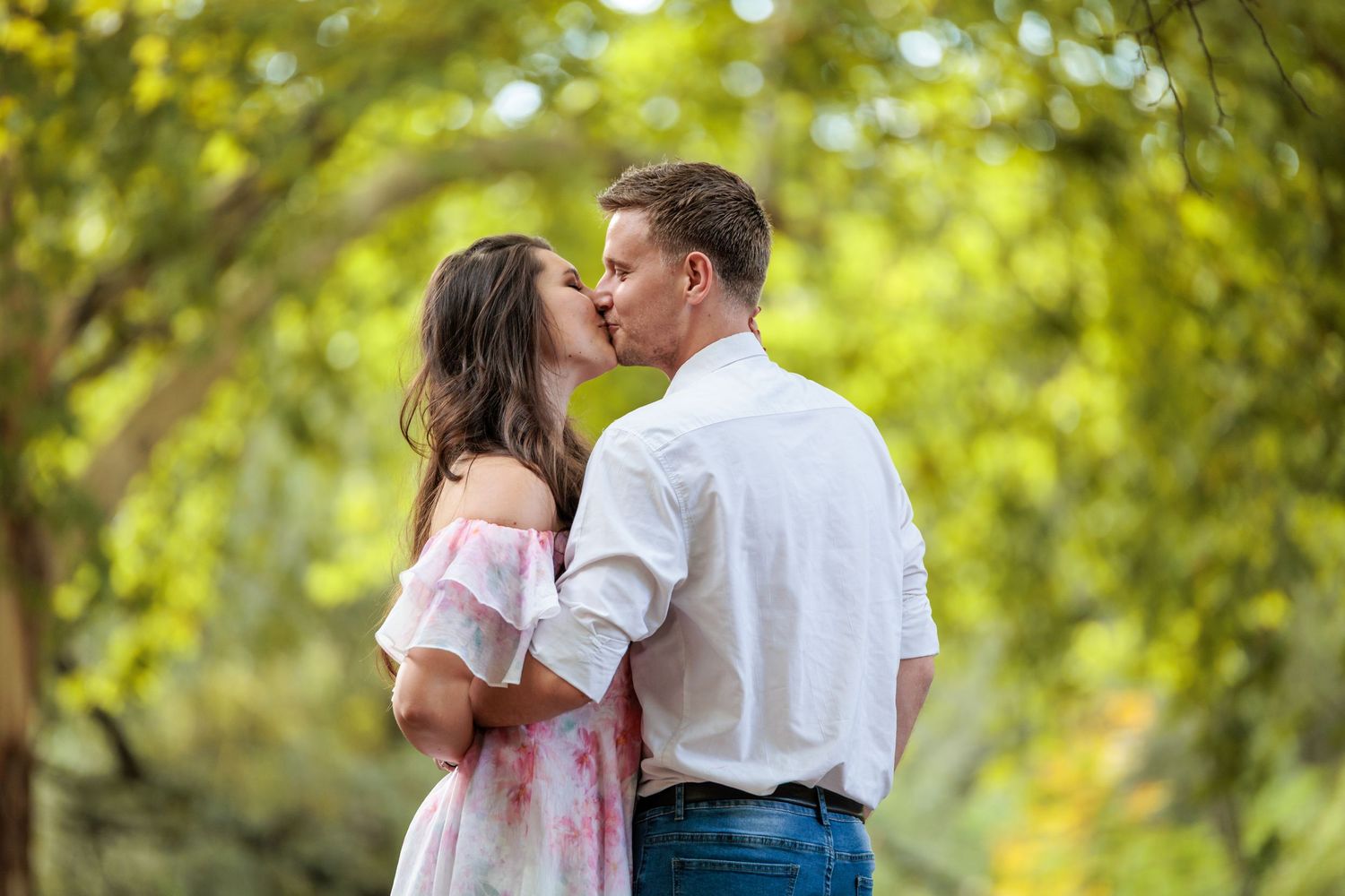 A romantic couple shares tender kisses in a sun-dappled outdoor setting with lush green foliage in the background.