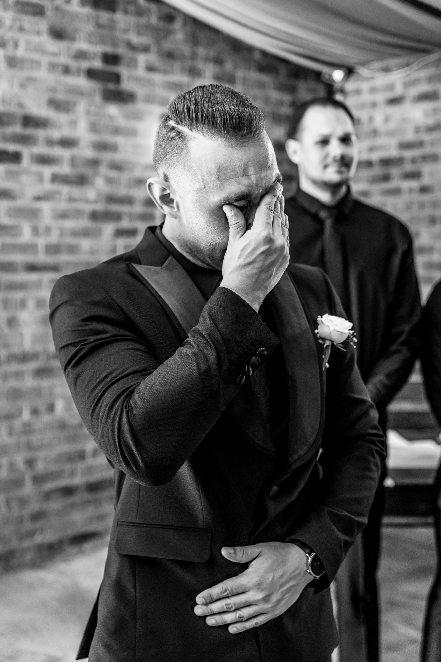 A groom in a black suit shows emotional reaction during wedding ceremony against brick wall backdrop.