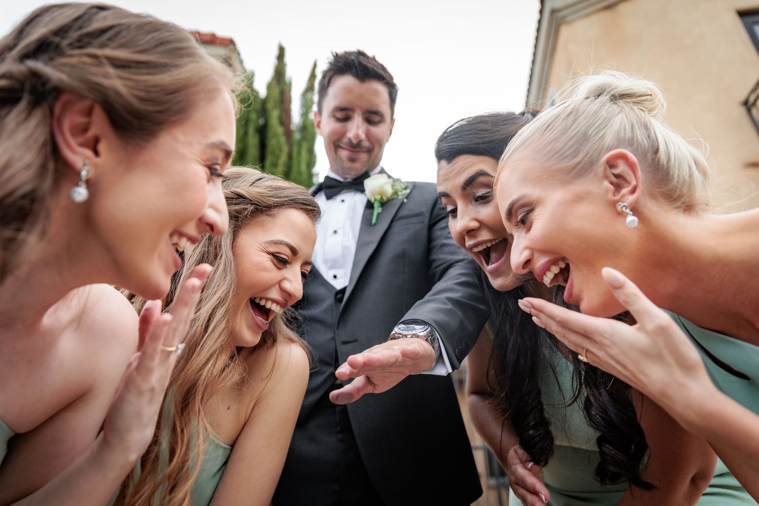 A group of wedding guests laugh together while huddling close and looking down at something.