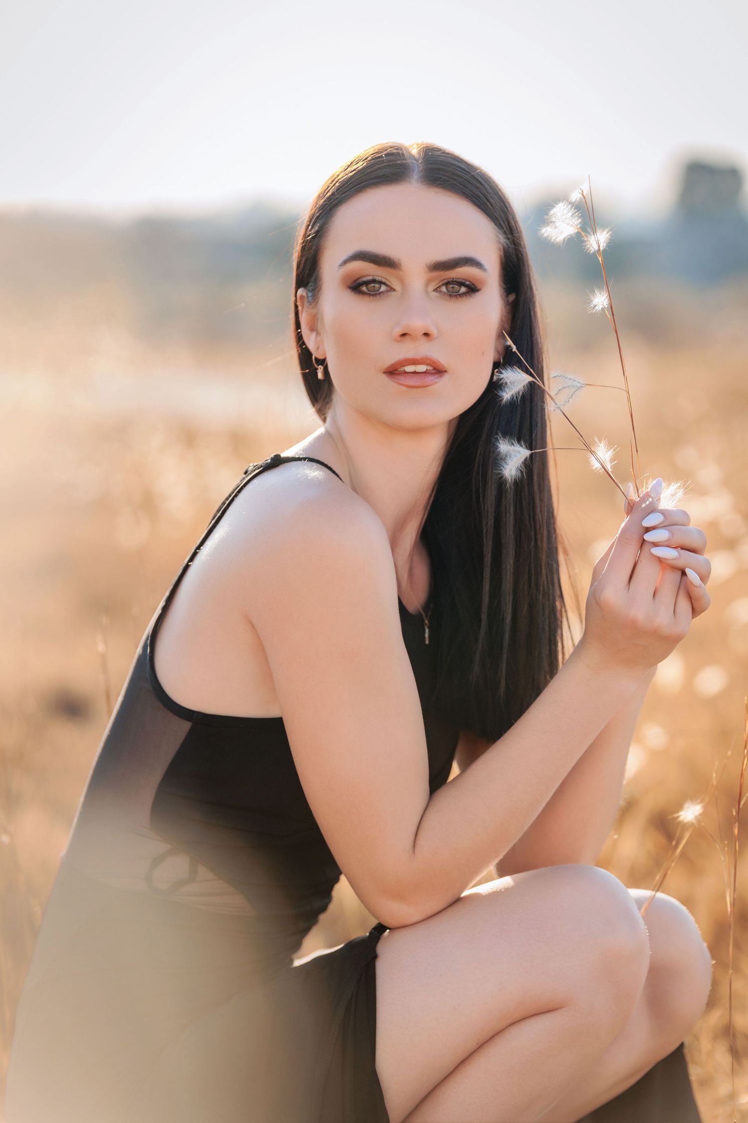 Portrait of a woman in a black dress posing in a golden field at sunset with dreamy bokeh lighting effects.