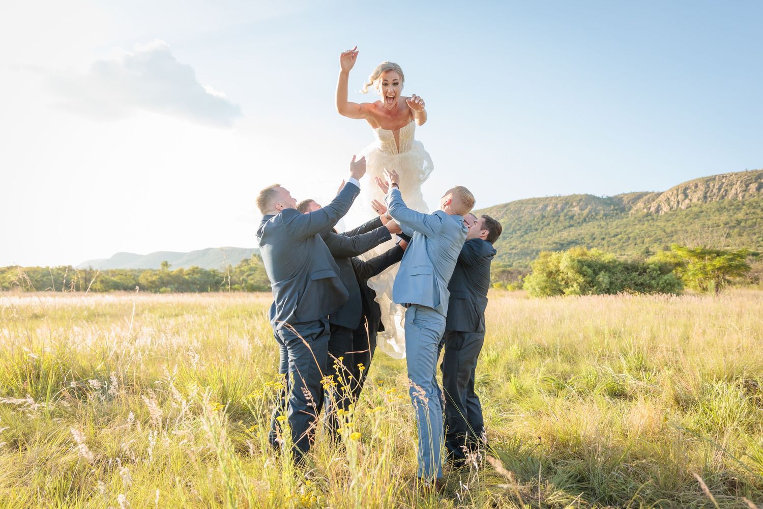 Bride lifted by groomsmen in sunny mountain meadow during outdoor wedding celebration.
