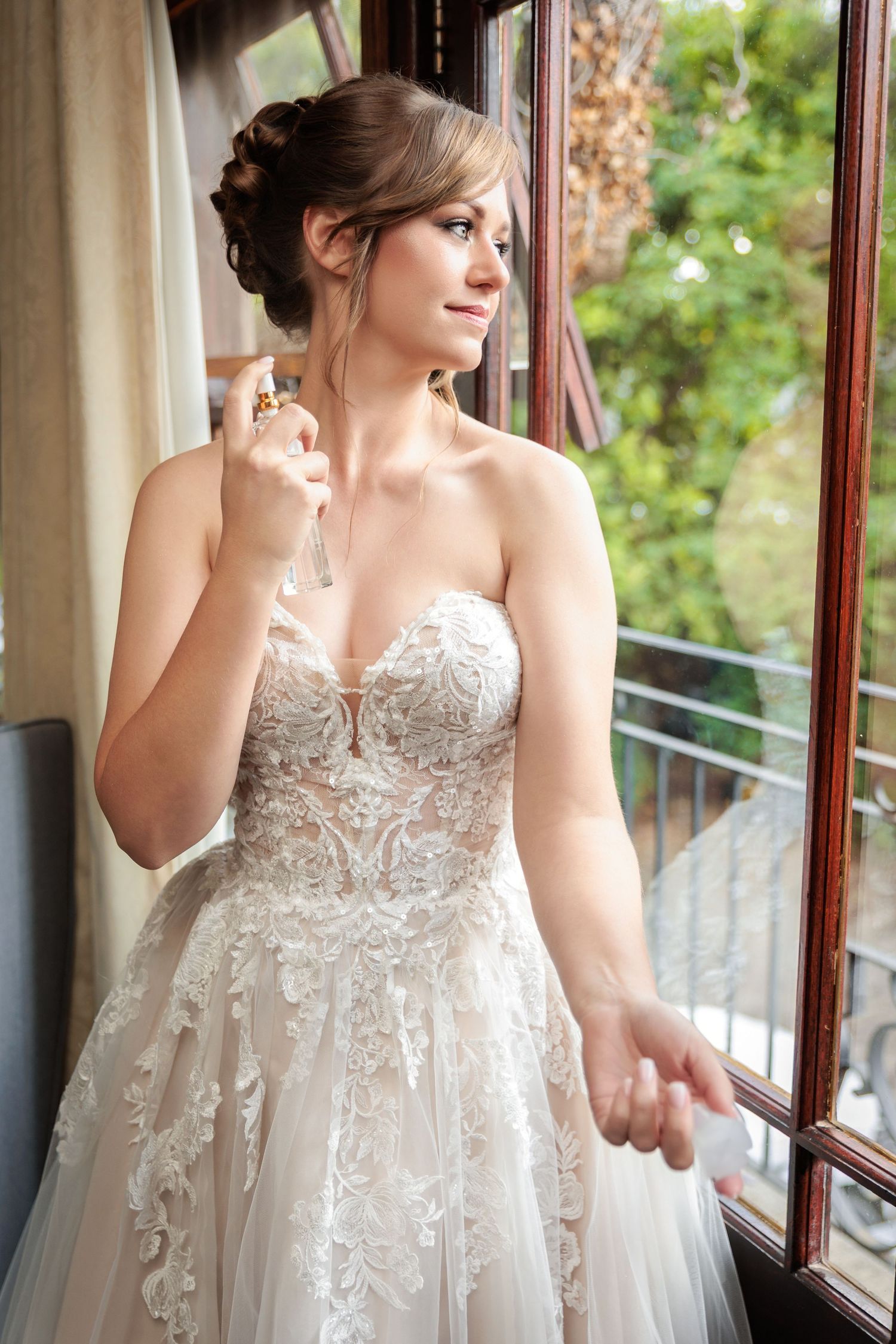 A bride in an elegant lace and tulle wedding gown stands near a window in a romantic setting.