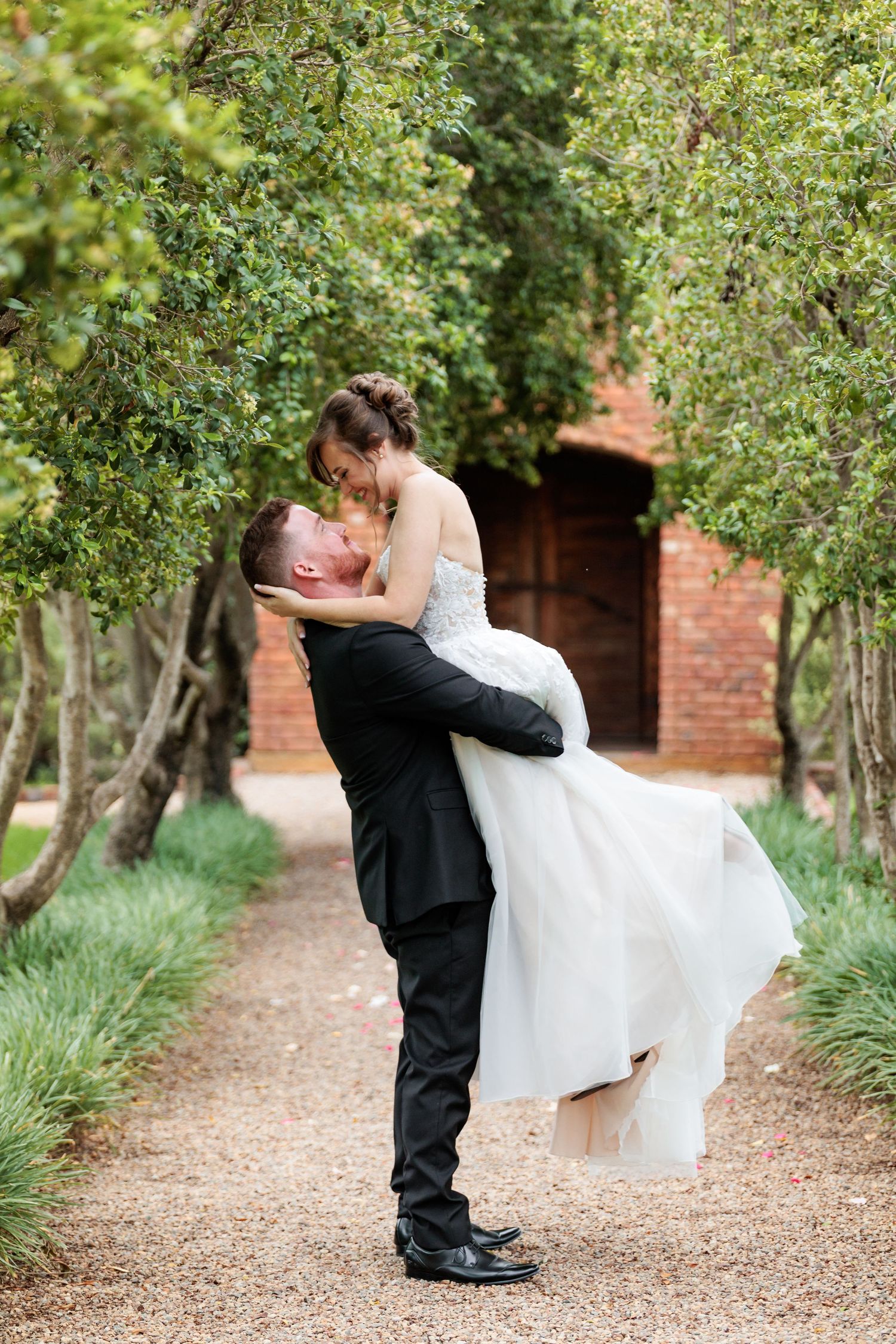 Romantic wedding couple embraces on a tree-lined path with brick building in background.