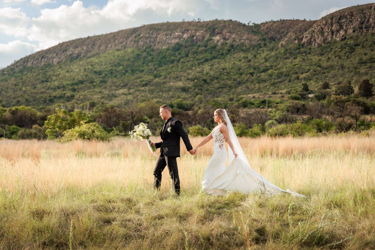 A couple in wedding attire runs playfully through a golden field with mountains in the background at sunset.