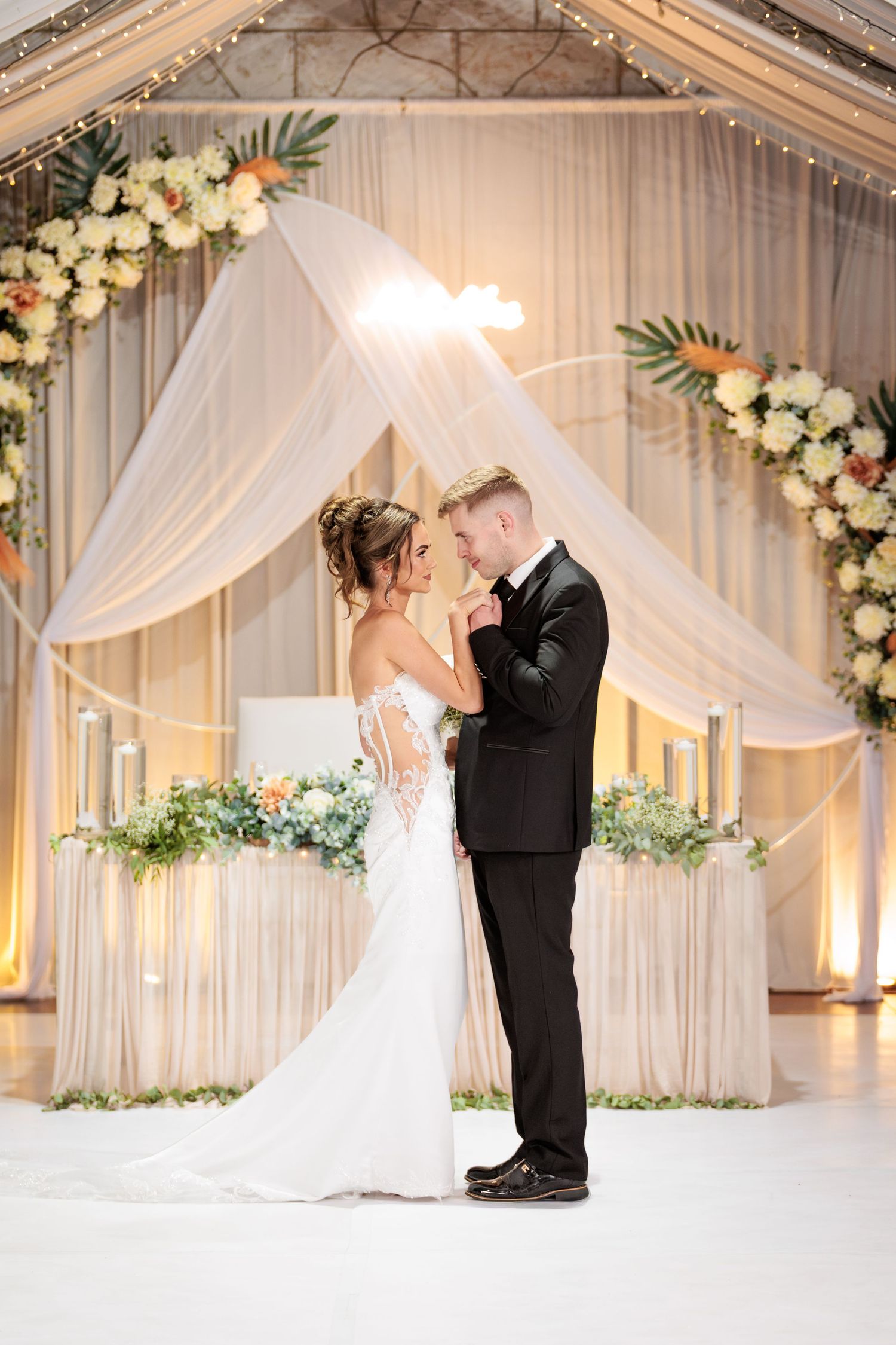 A romantic wedding ceremony setup with white draping, floral arch decorations and soft lighting creating an elegant atmosphere.