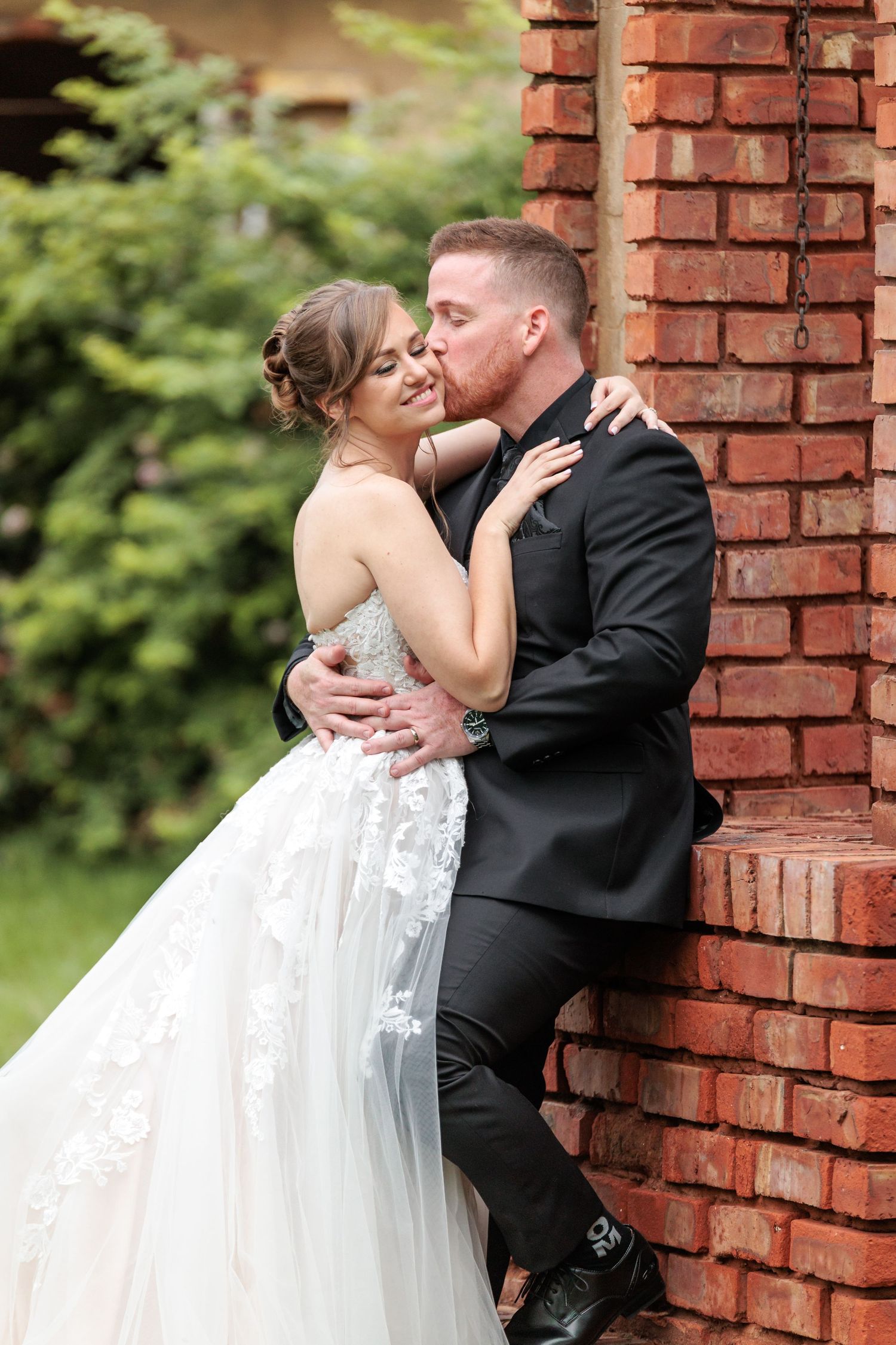 A romantic wedding couple shares intimate kisses against a rustic brick wall in a garden setting.