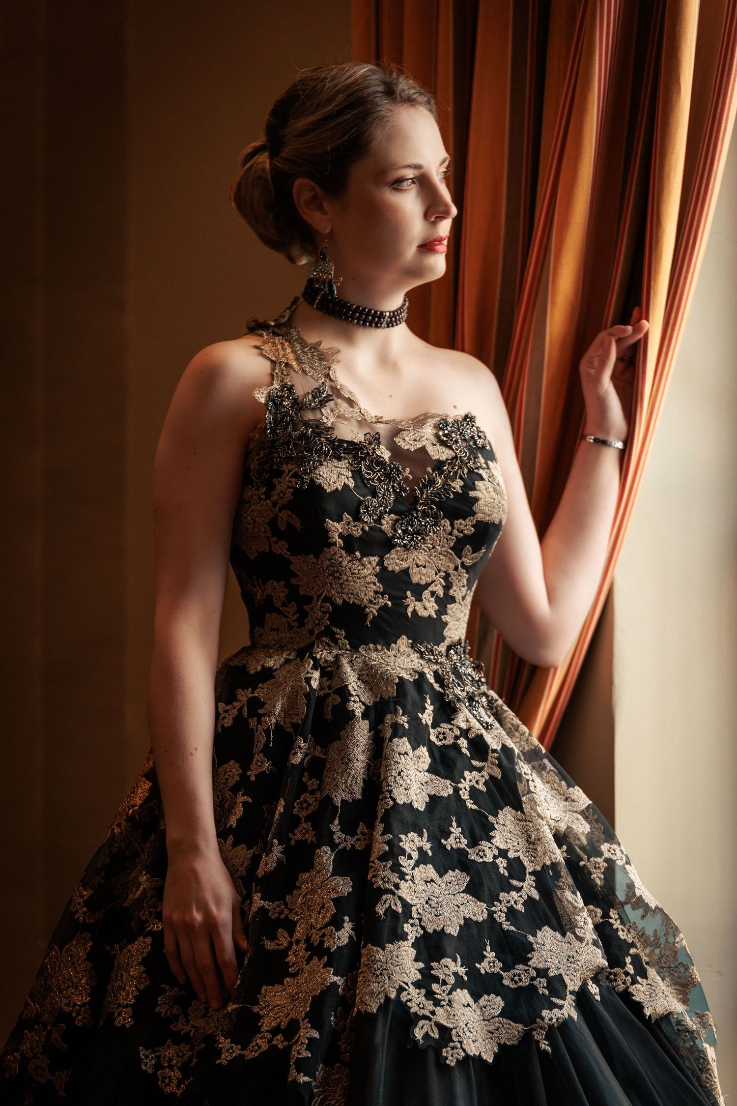 A model poses in a dramatic black and gold lace evening gown with floral appliques against a warm-toned interior.