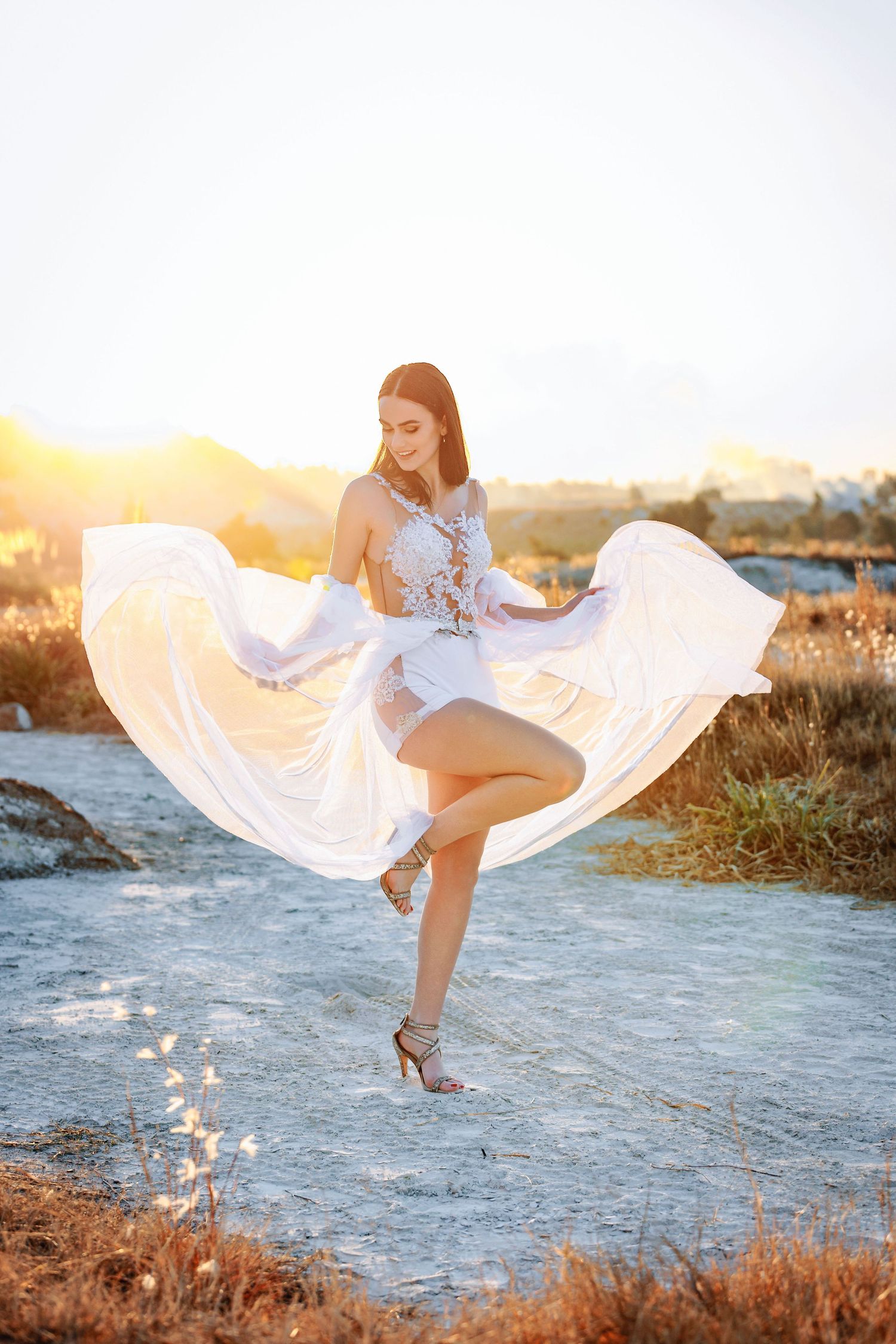 A white flowing dress billows in the breeze as someone poses gracefully near the ocean at sunset.